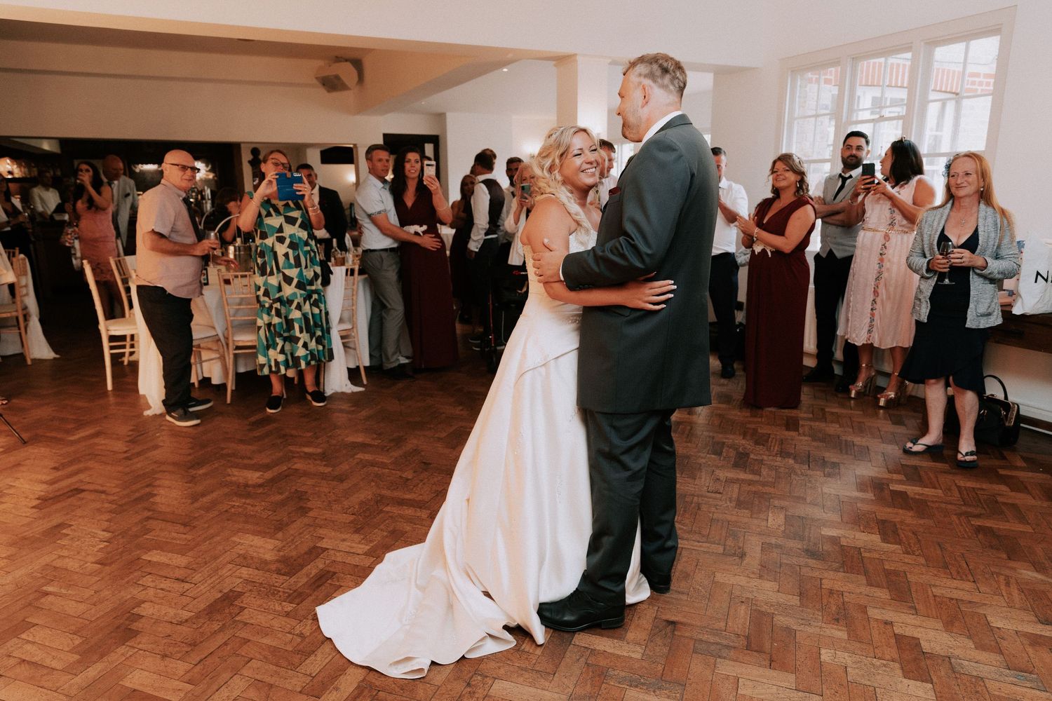Bride and groom share their first dance on a wooden floor surrounded by wedding guests at a elegant reception venue.