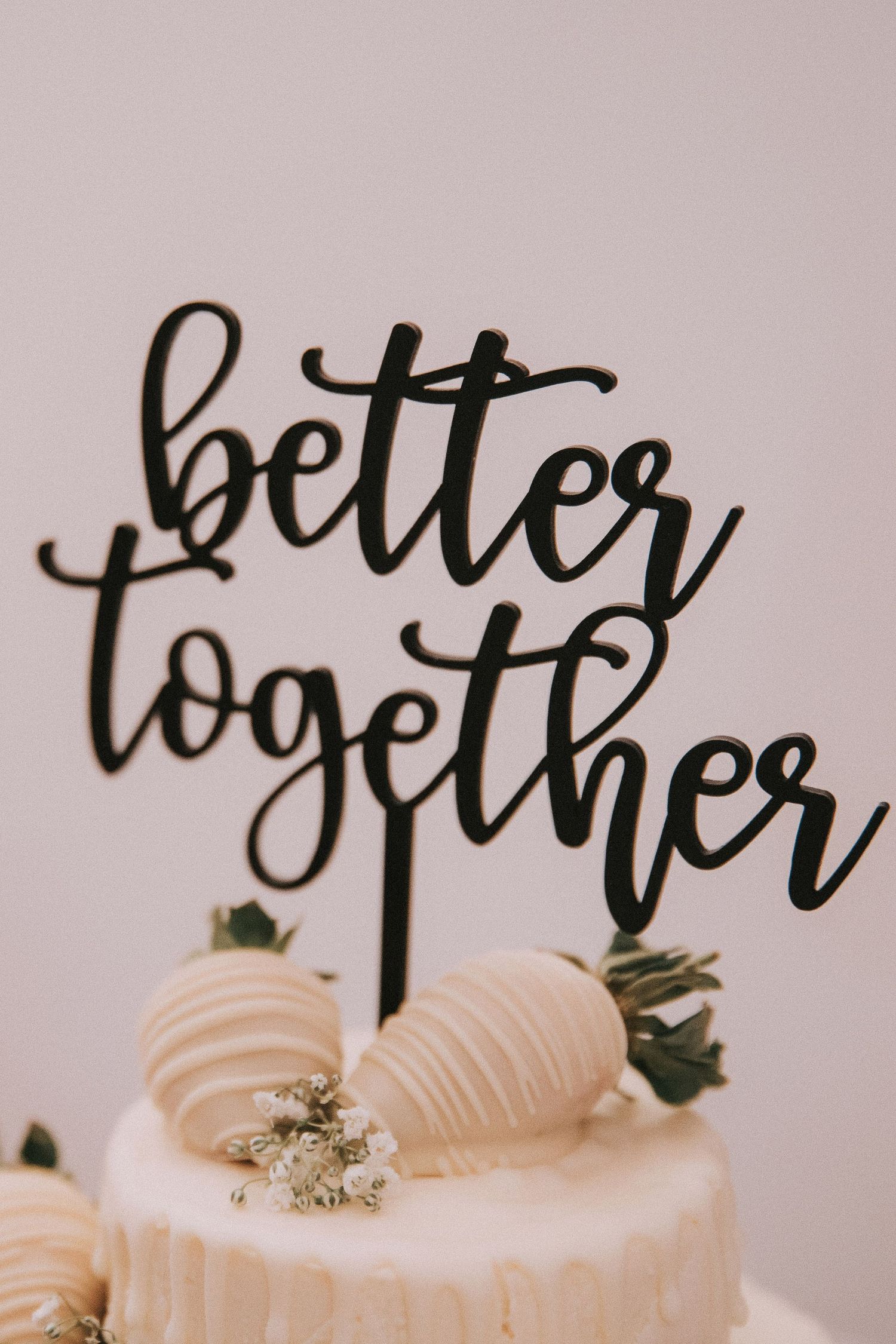 Black laser-cut cake topper reading better together on a white wedding cake with strawberries.