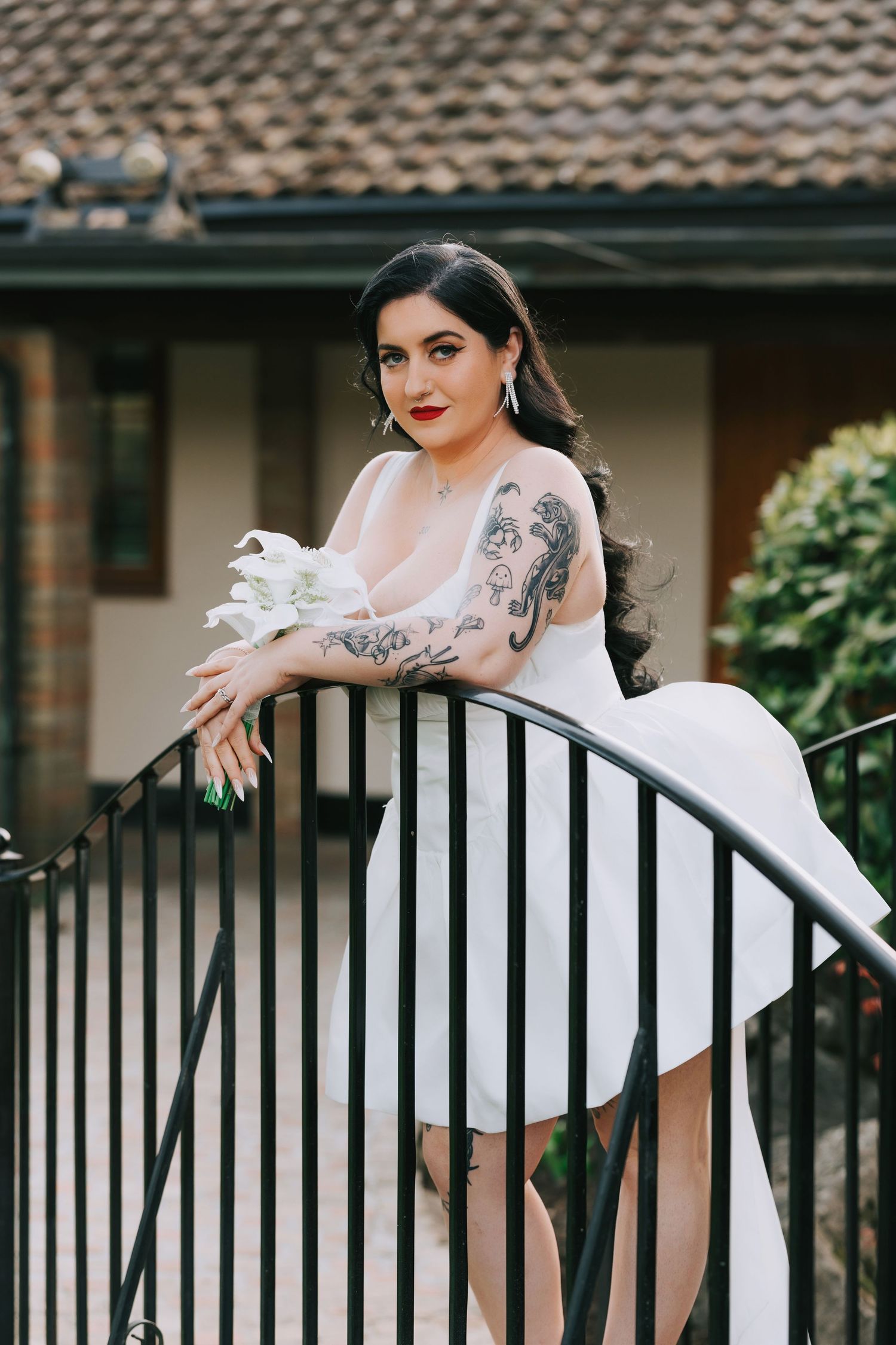 Tattooed bride in white dress leans on black iron railing holding white flower, red lips, outdoor venue.