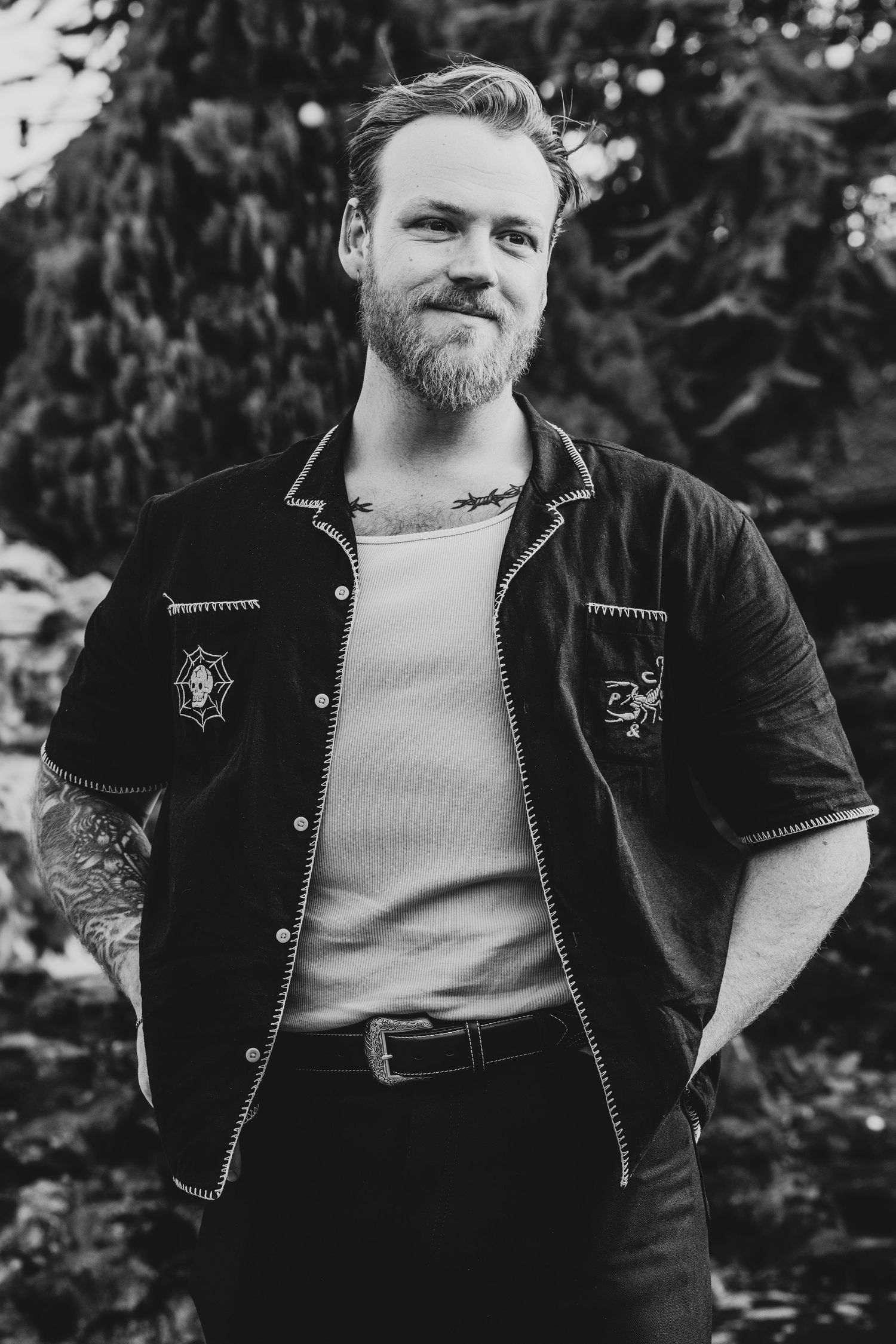 Black and white portrait of a bearded tattooed man smiling outdoors wearing a dark embroidered shirt.