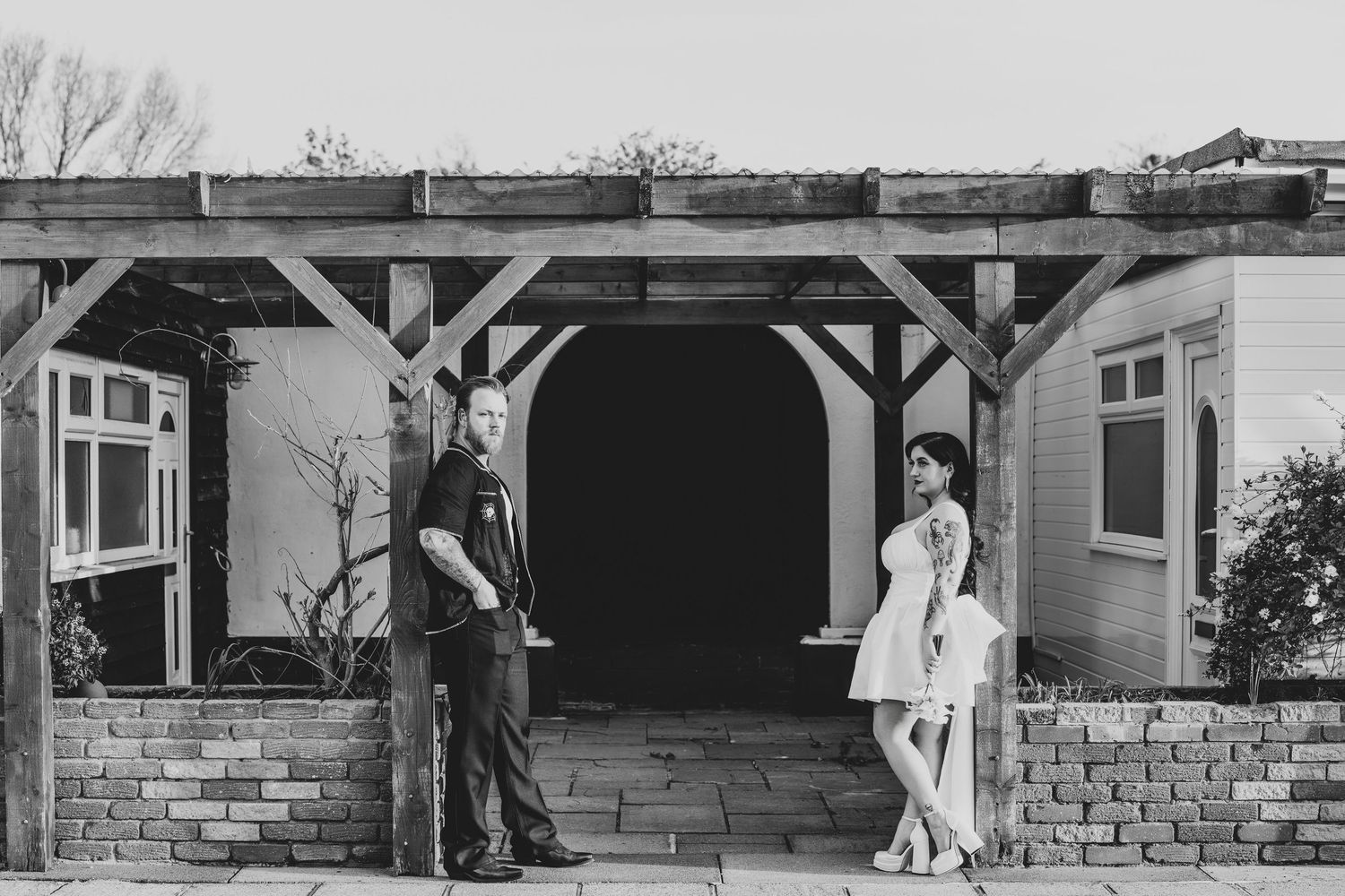 Black and white photo of a couple posing under a wooden pergola outside a rustic building on a paved patio.