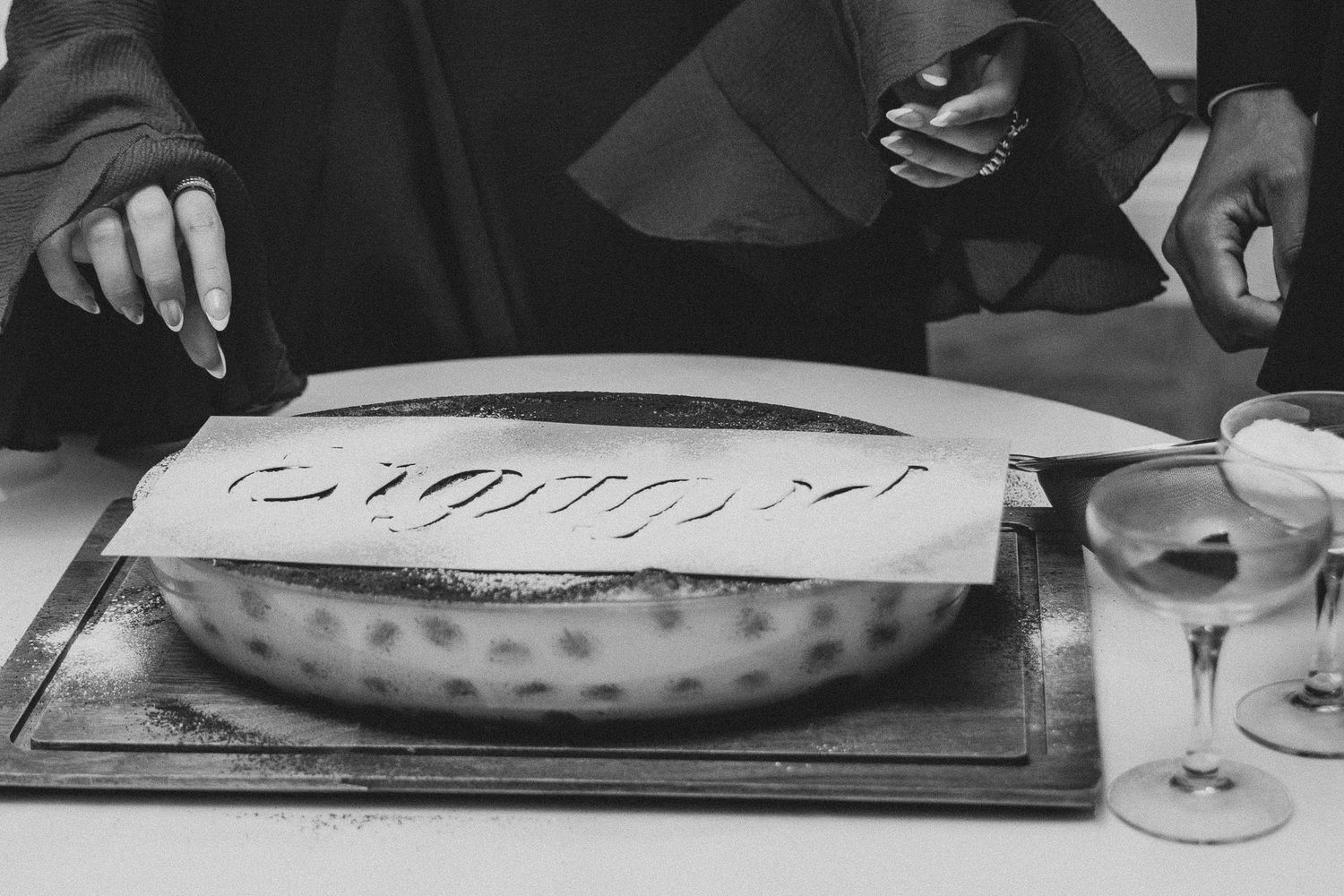 Black and white photo of a person cutting an elegantly decorated oval cake on a tray beside a glass.