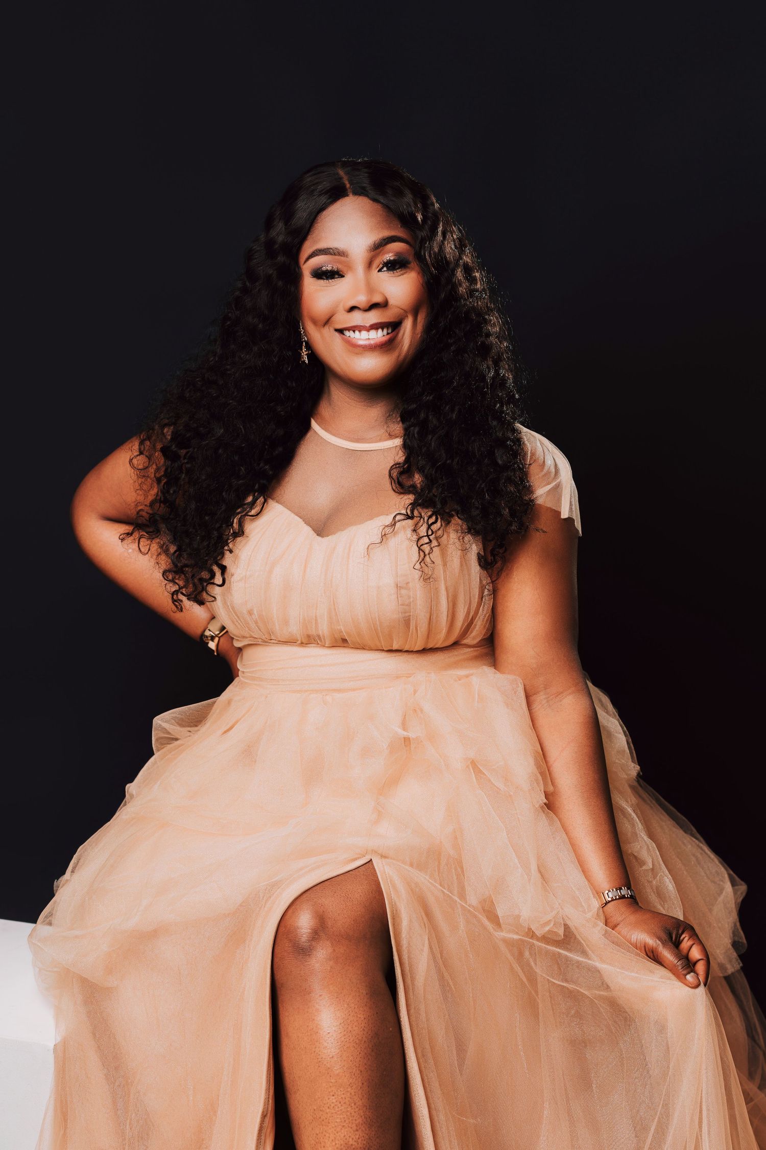 Smiling woman in elegant blush tulle gown with curly hair poses confidently against dark background.