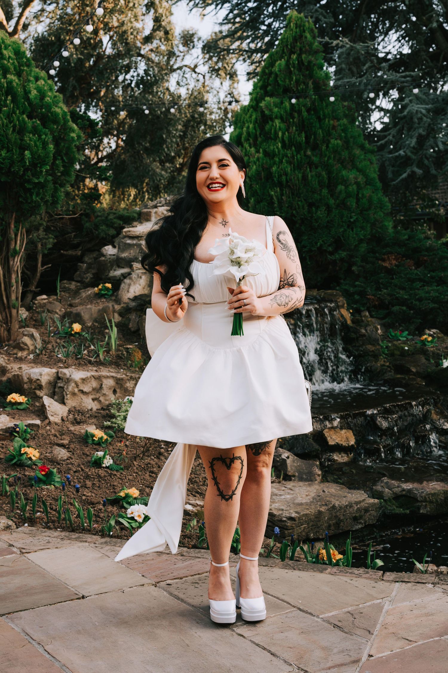 Tattooed bride in short white dress holding bouquet, standing in garden with waterfall backdrop.
