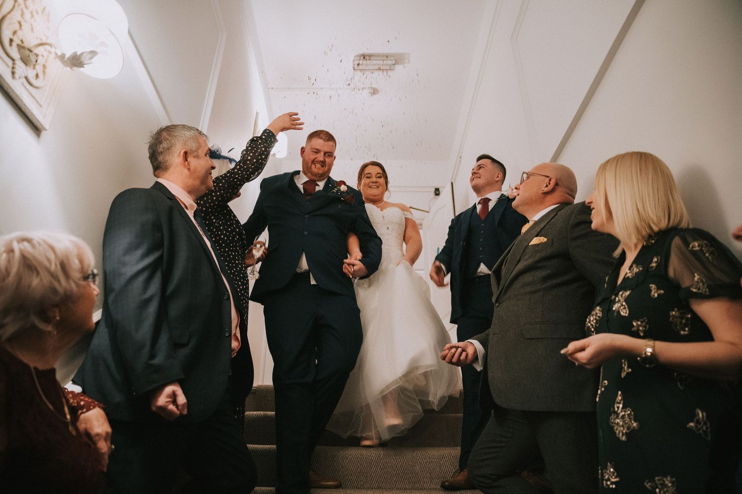 Joyful newlyweds celebrate with family and friends in a hallway, guests throwing confetti after the wedding.