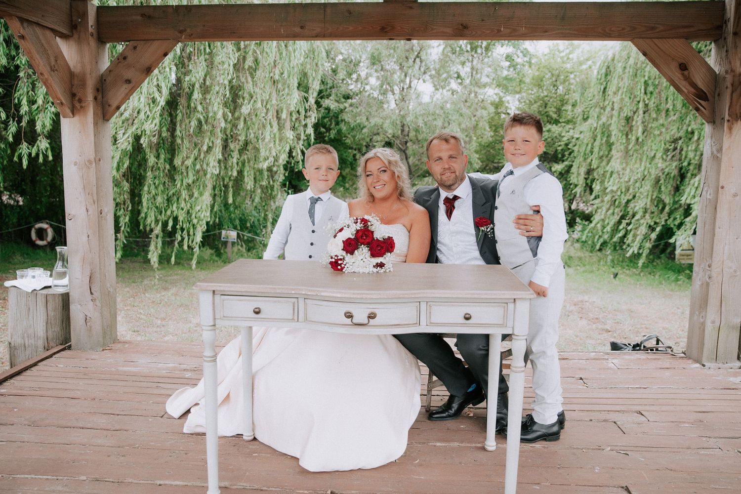 Bride and groom pose with two boys in white suits at a rustic outdoor wedding ceremony under a wooden pergola.