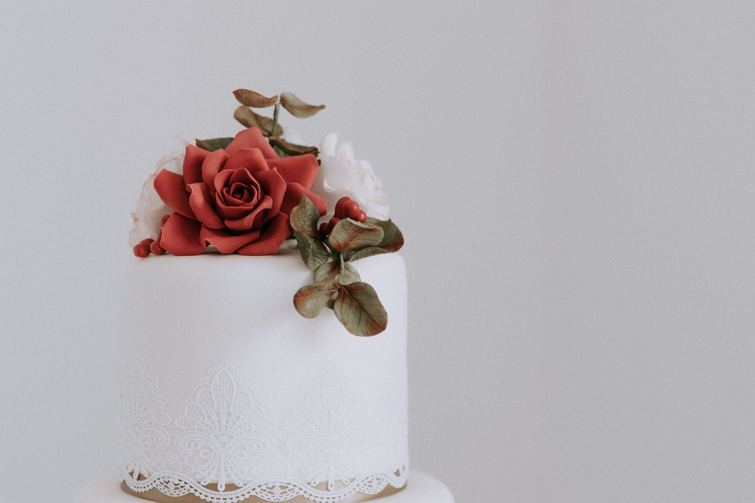 White two-tier wedding cake topped with red roses, white flowers, and eucalyptus leaves on a light gray background.