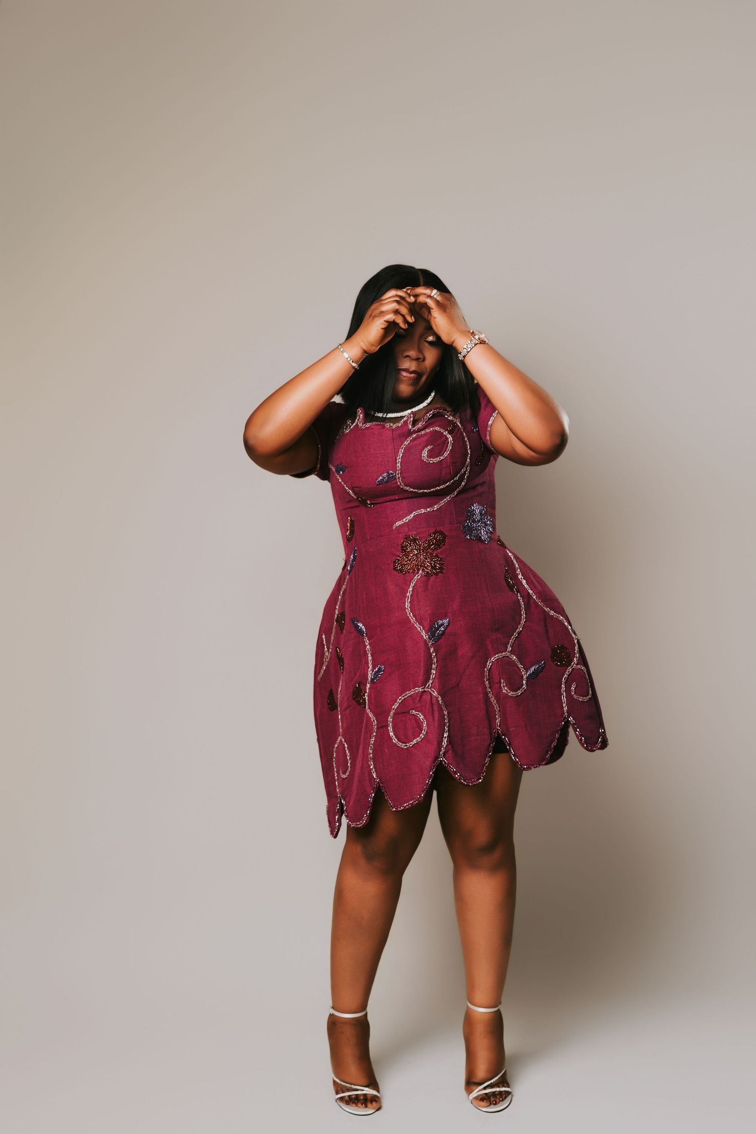 Woman in burgundy beaded mini dress with scalloped hem and white heels posing with hands on head.