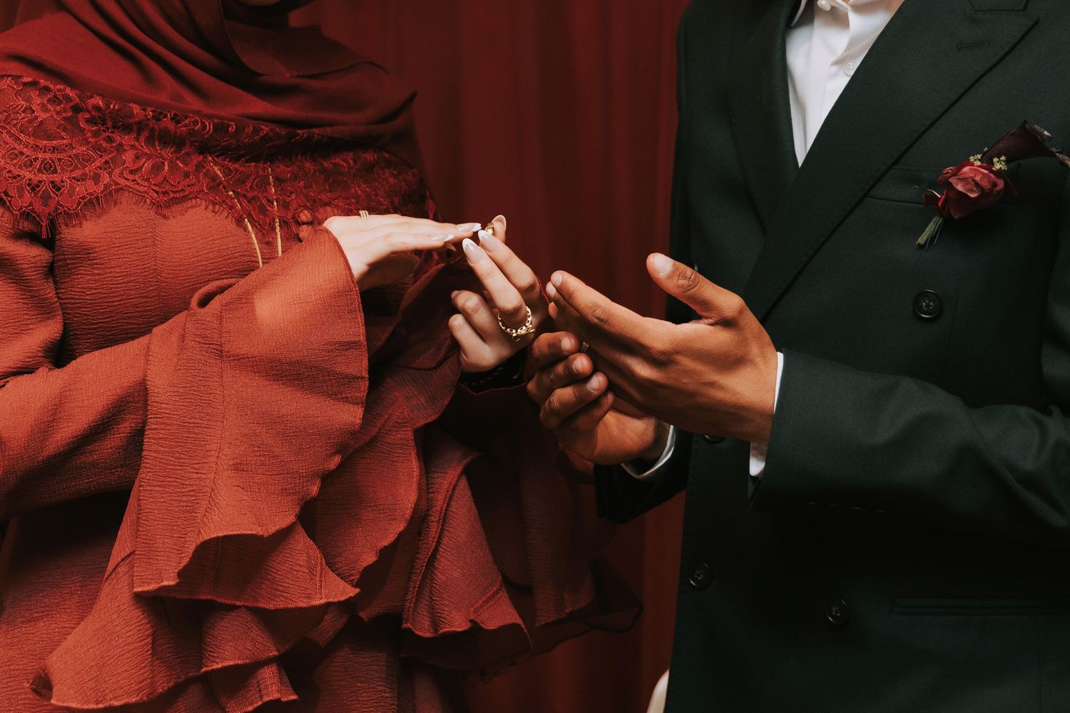 A groom in a black suit places a ring on a bride's finger as she wears a red lace dress at a wedding ceremony.