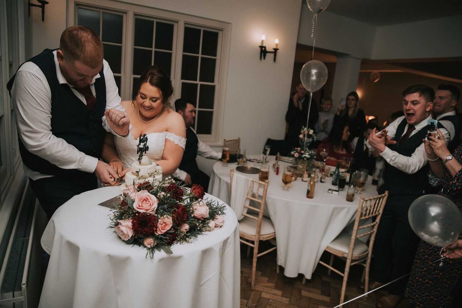 Bride and groom cut their wedding cake together at a decorated reception table with floral centerpiece.