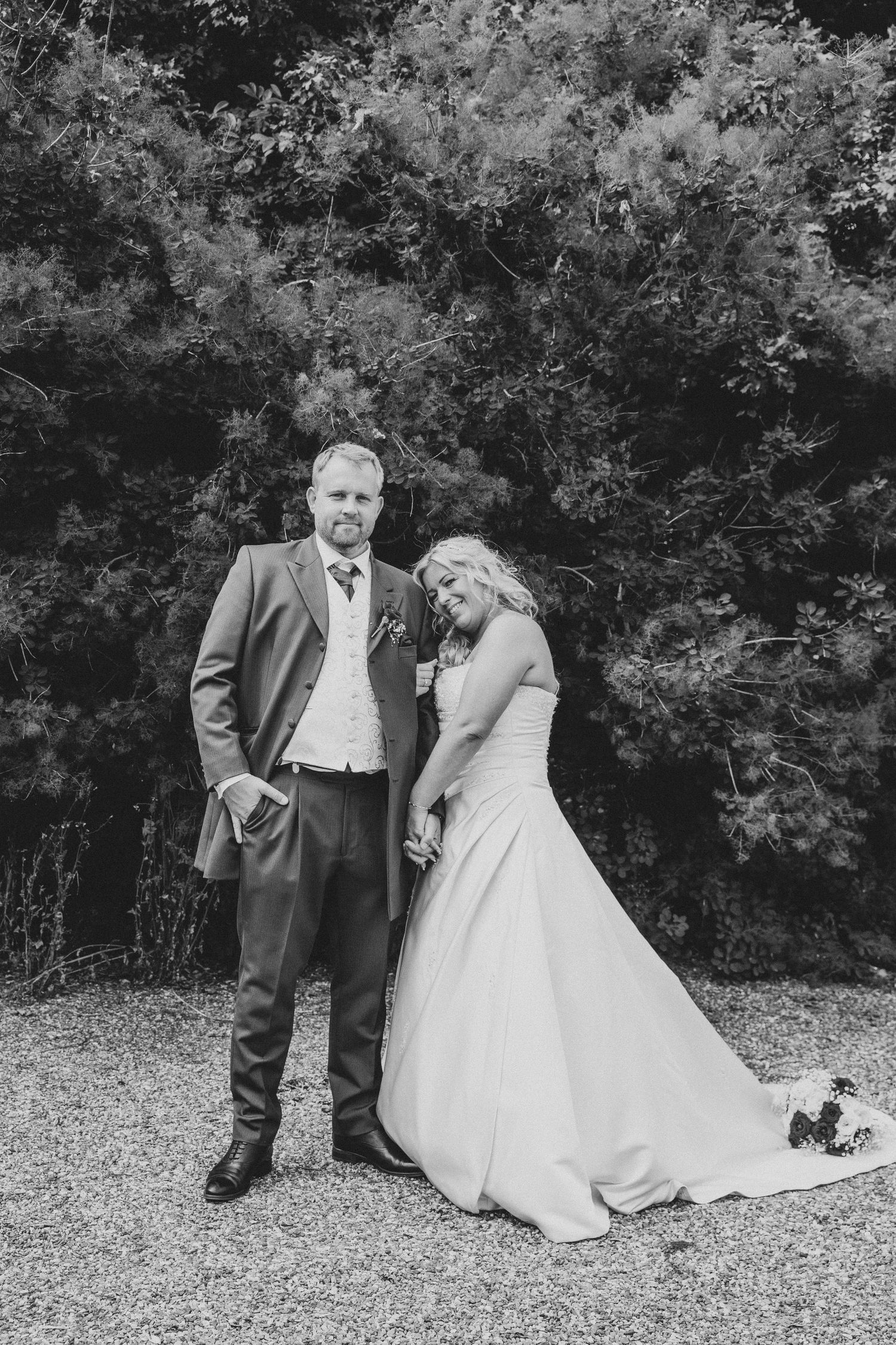Black and white portrait of bride and groom holding hands before lush green trees outdoors.