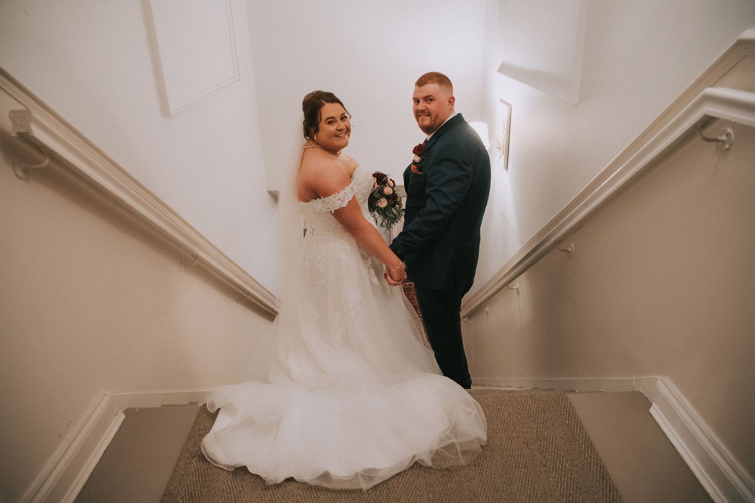 Bride and groom holding hands on staircase, bride in off-shoulder white gown, groom in navy suit, smiling at camera.