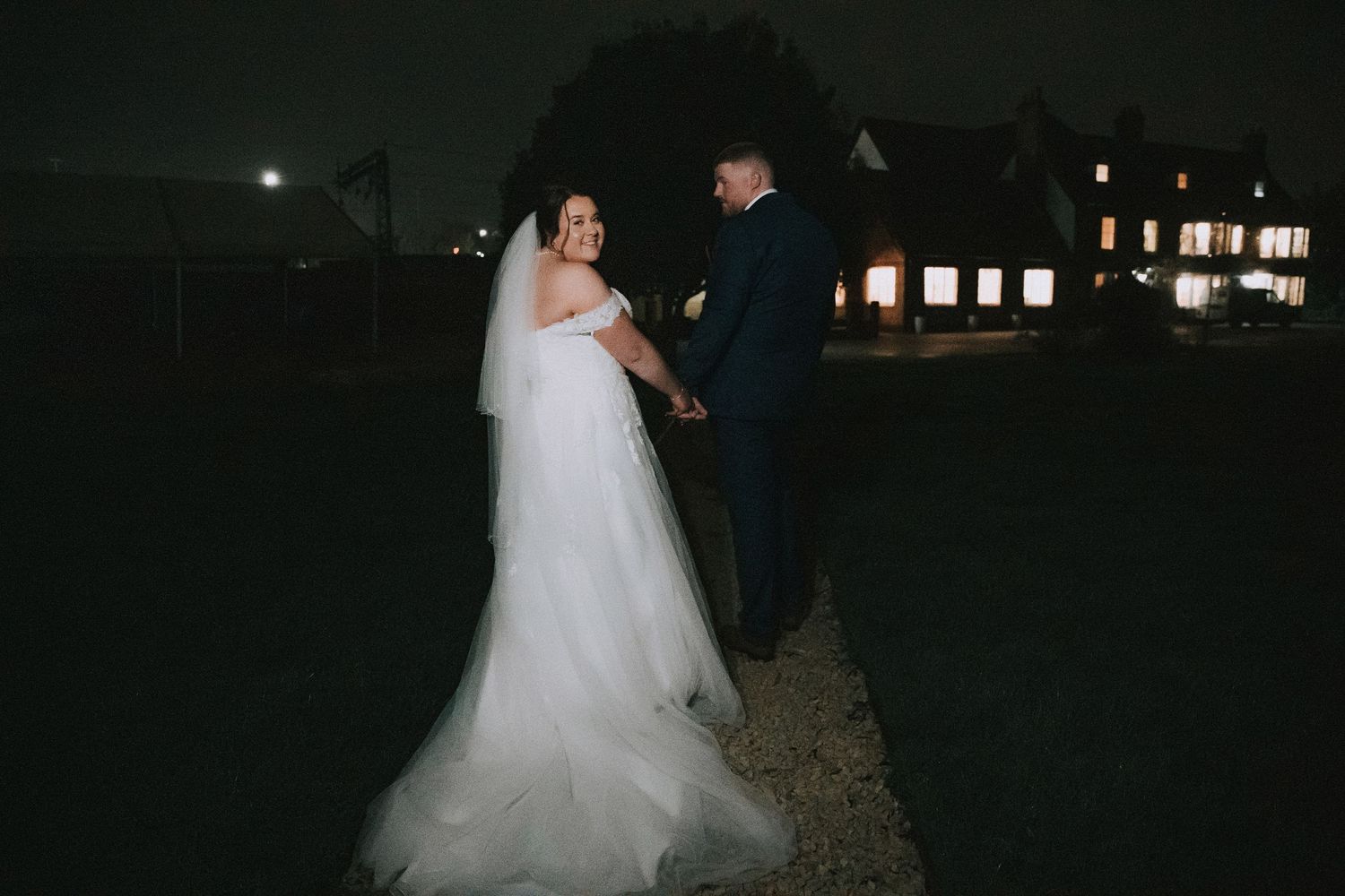 Bride and groom holding hands outdoors at night with a lit venue building in the background.