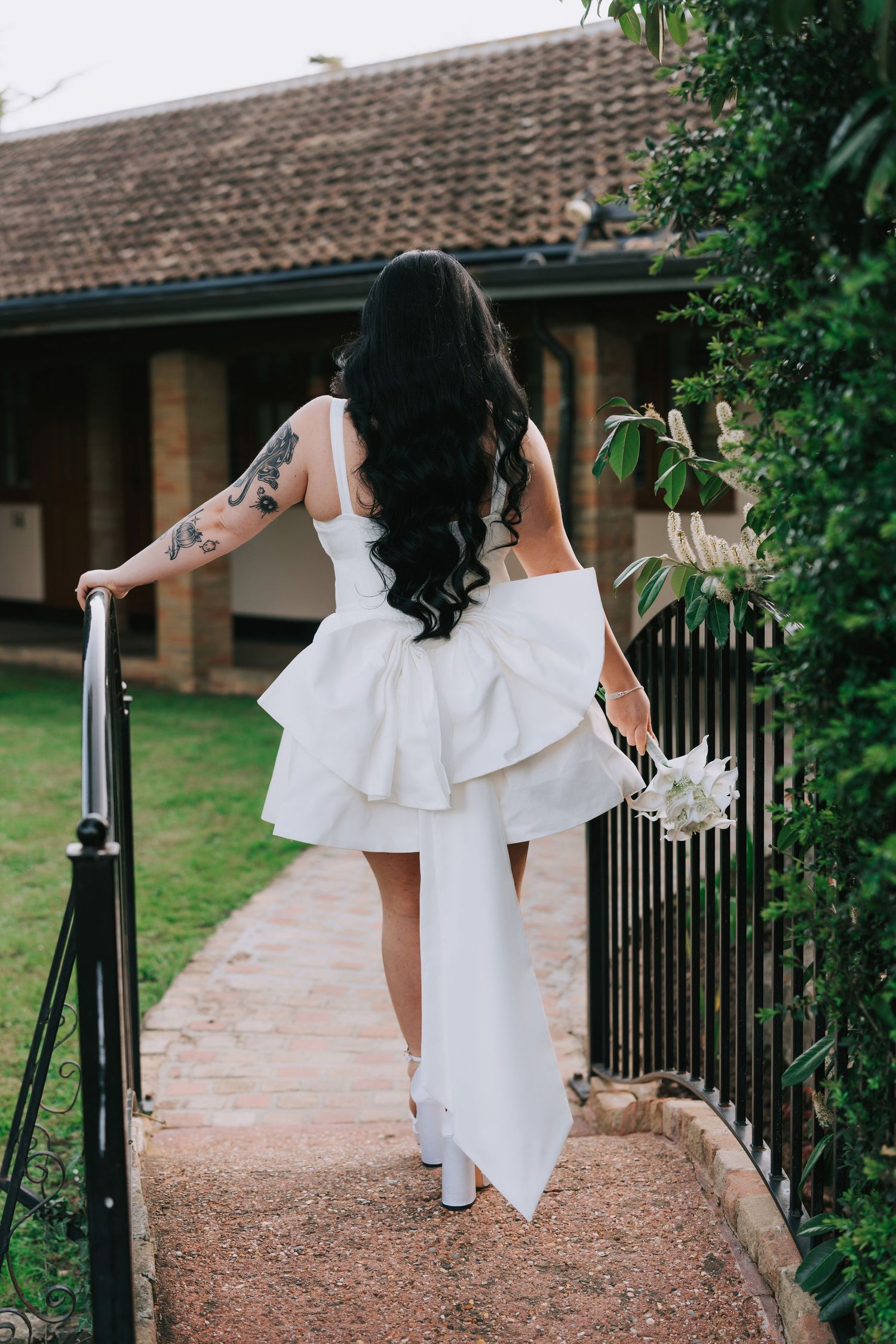 Bride in white hi-lo dress with tattoos holds bouquet, walking along the brick path beside iron fence at Ye Olde Plough House Essex