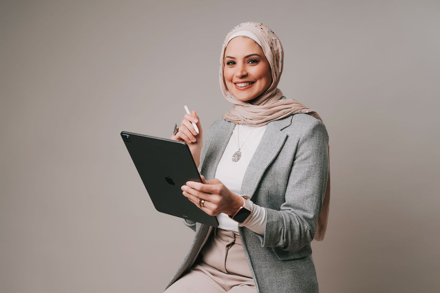 Professional headshot of a woman in hijab holding tablet and pointing, wearing gray blazer, smiling confidently at Basildon Studio
