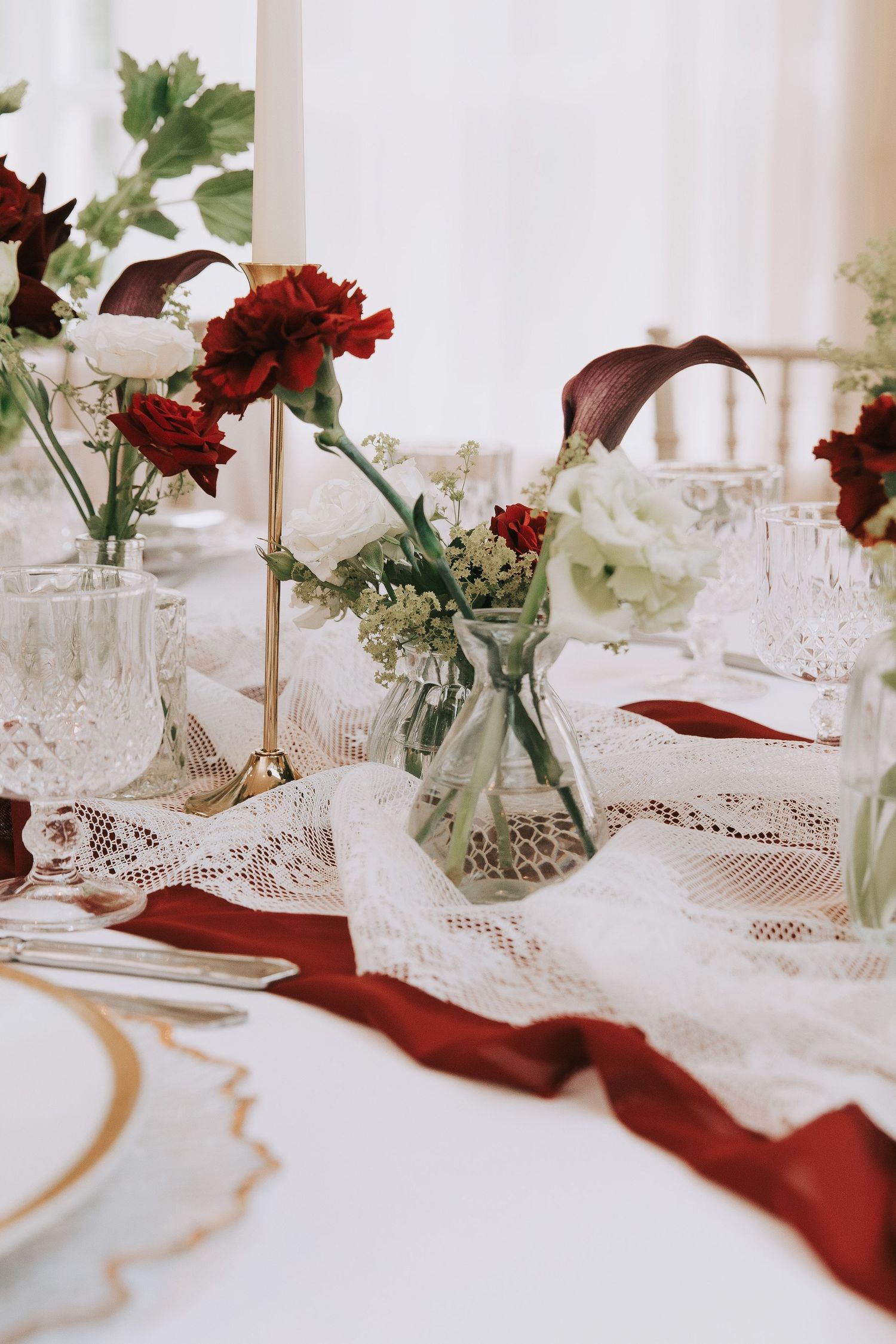 Elegant wedding table setting with red roses, white lace runner, crystal glasses, and crimson napkins.