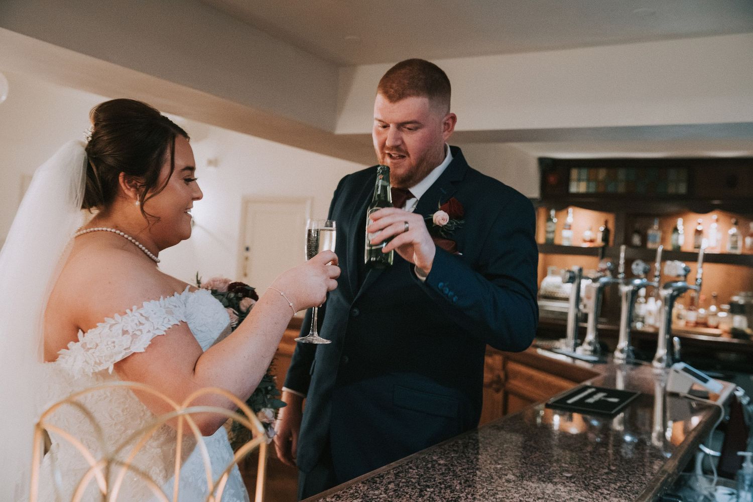 Bride and groom in navy suit share a champagne toast at a bar during their wedding reception.