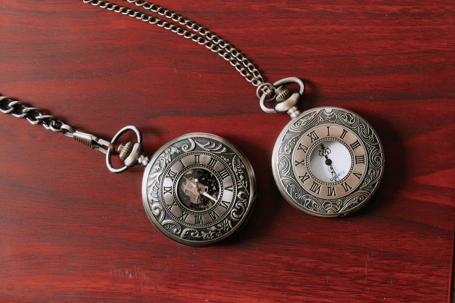Two antique bronze pocket watches with Roman numerals on a rich red wooden surface, connected by a decorative chain.