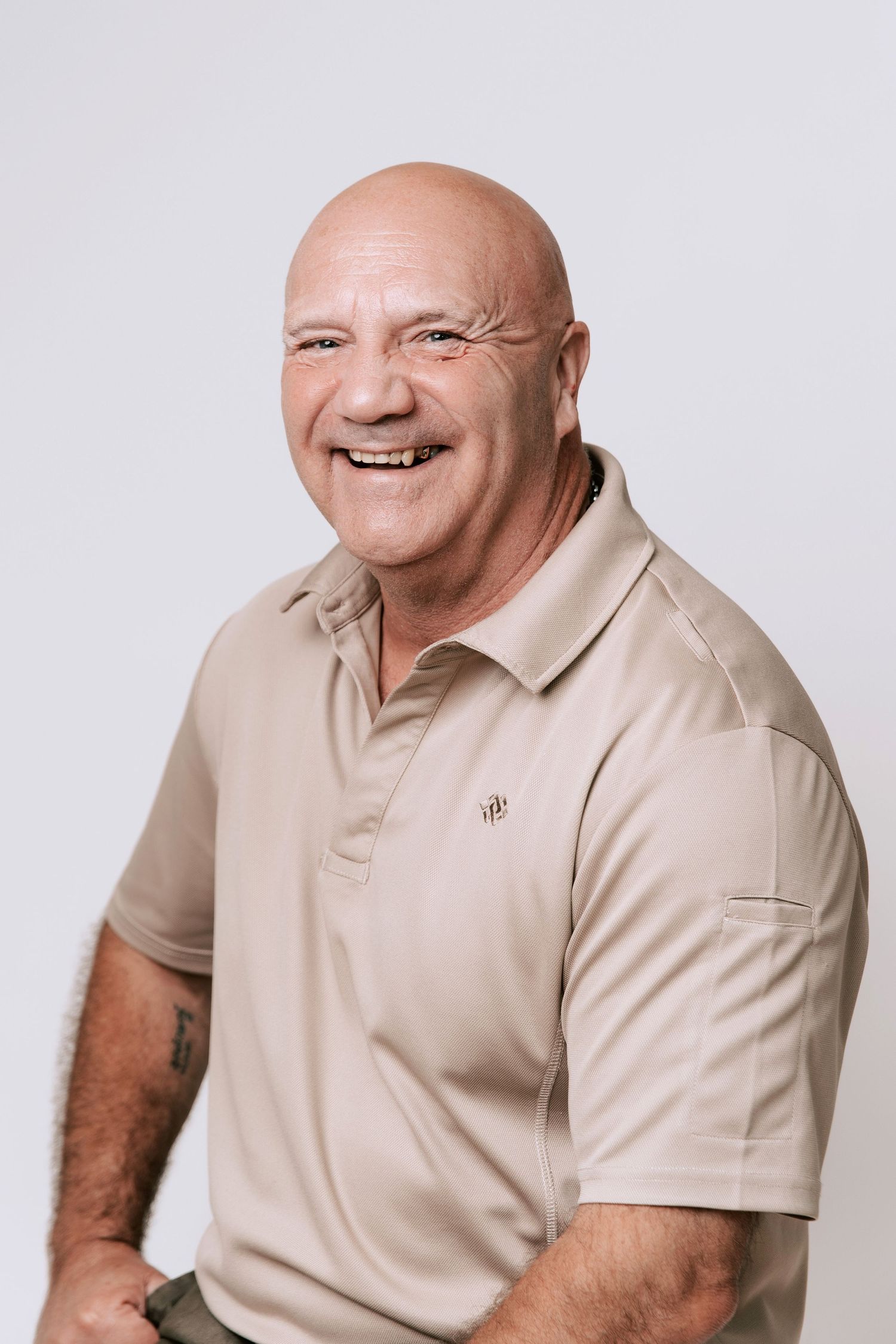 A man wearing a beige polo shirt is sitting and smiling at the camera against a plain light background, ideal for professional headshots