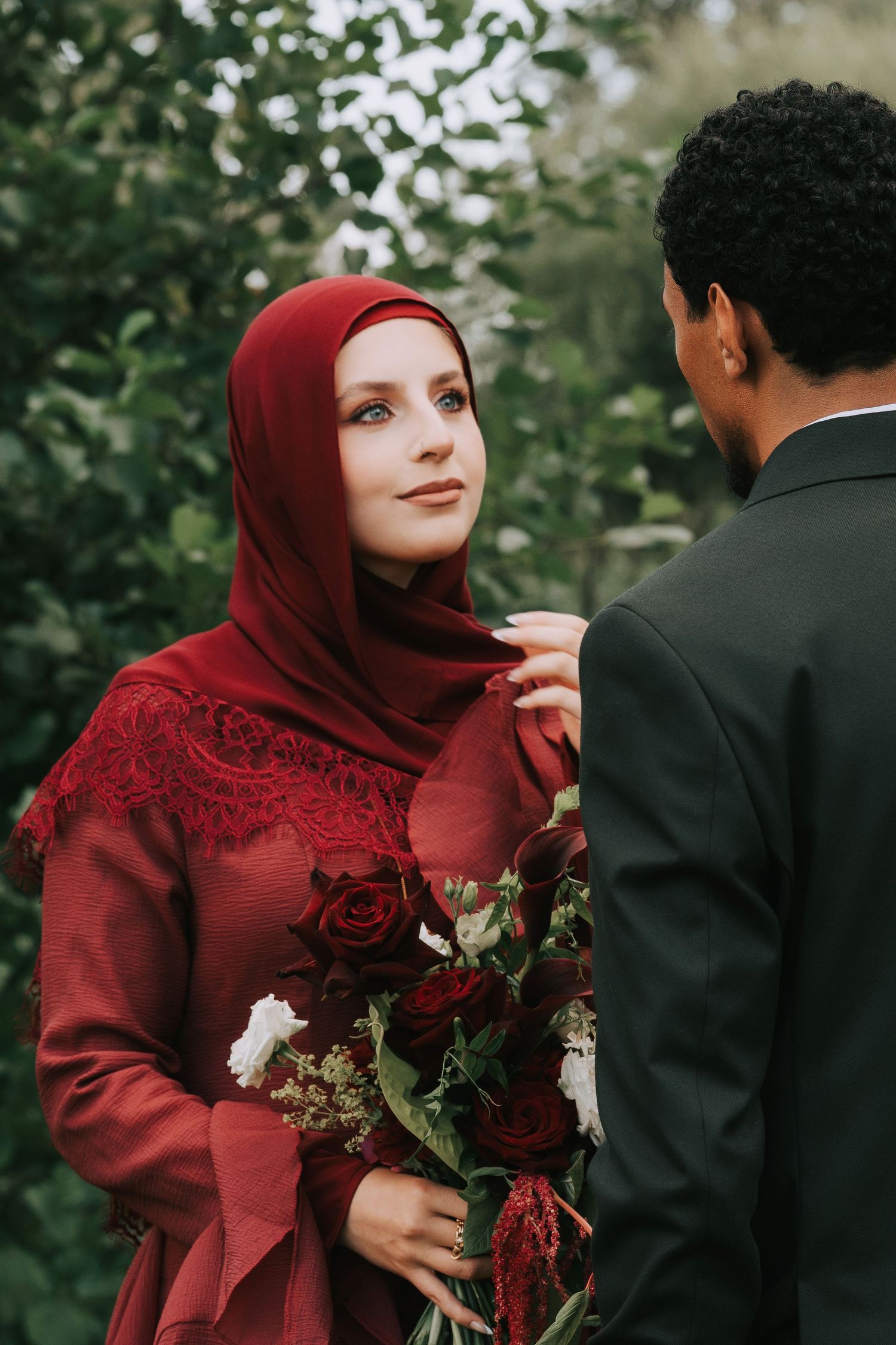 A woman in a red hijab and floral dress holds roses while gazing at a man in a dark suit outdoors.