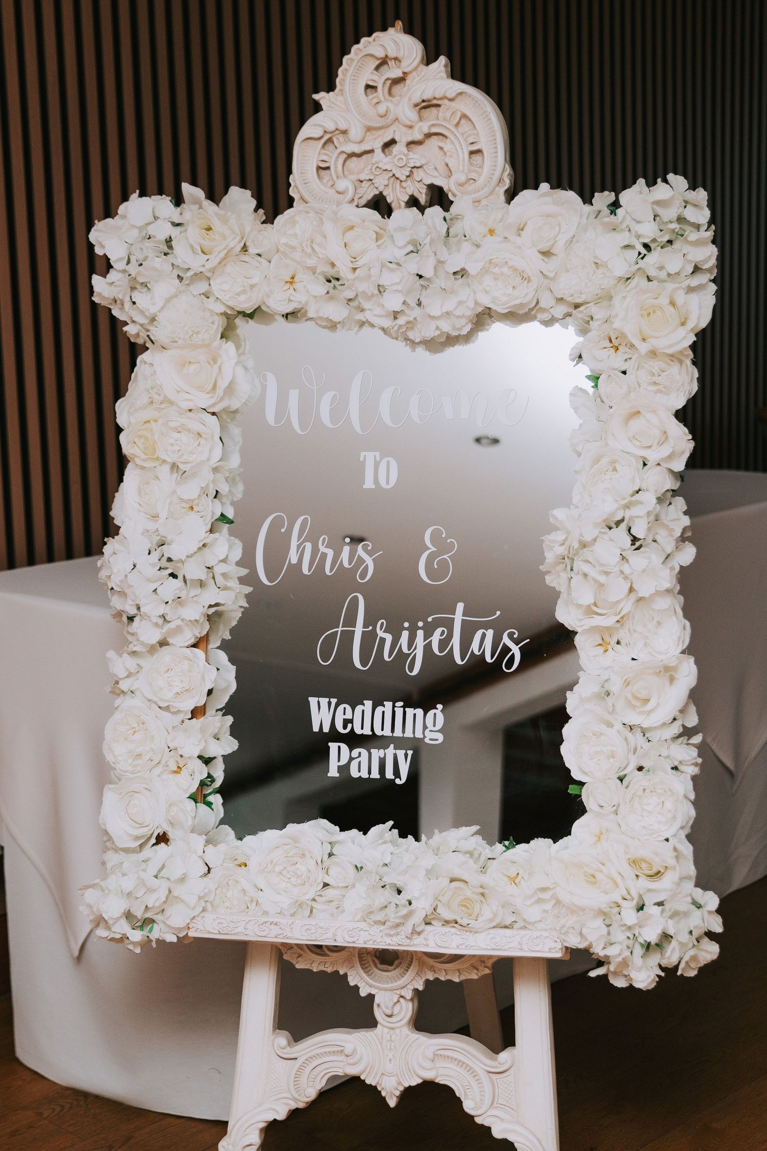 Elegant wedding welcome mirror sign with white floral frame reading Welcome to Chris and Arijetas Wedding Party.