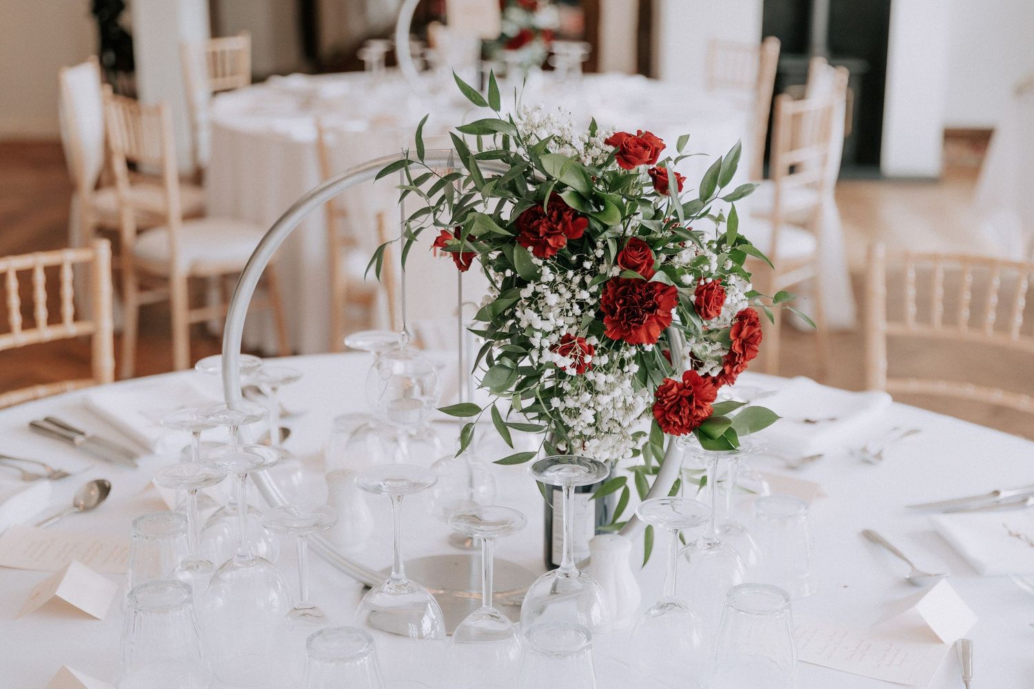 Elegant wedding table centerpiece with red roses, greenery, and baby's breath on a white tablecloth with glassware.