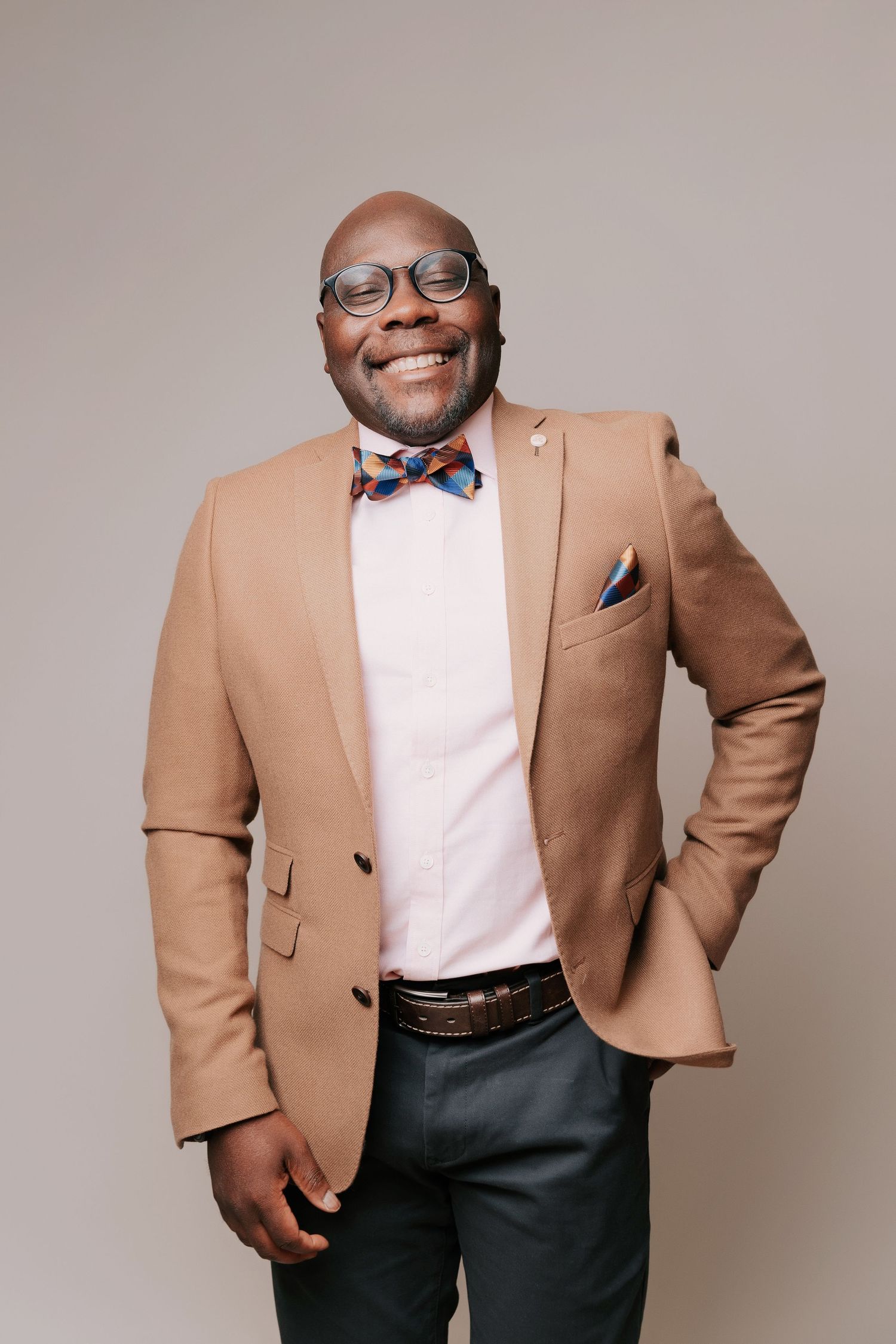 A smiling man in glasses, a tan blazer and colorful bow tie, and dark pants creating a polished look perfect for professional headshot photo