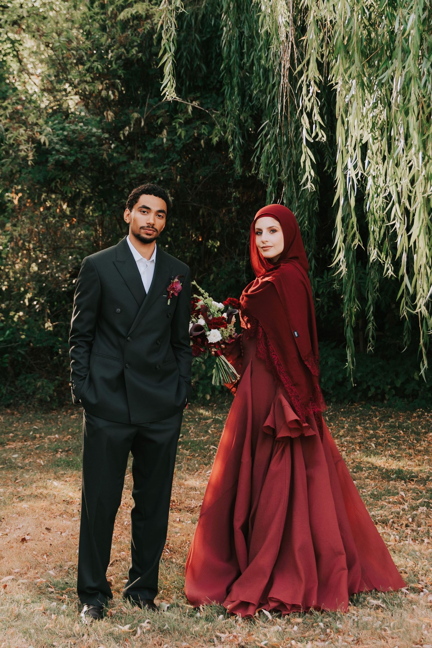 Couple in wedding attire, man in black suit, woman in red hijab gown, posing outdoors near weeping willow.