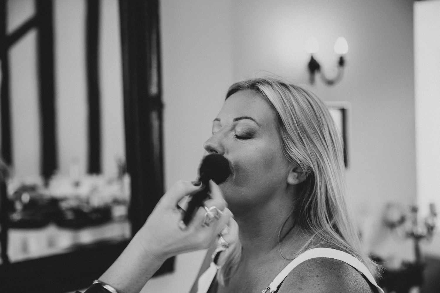 Black and white photo of a blonde woman applying makeup with a brush in an elegant room.
