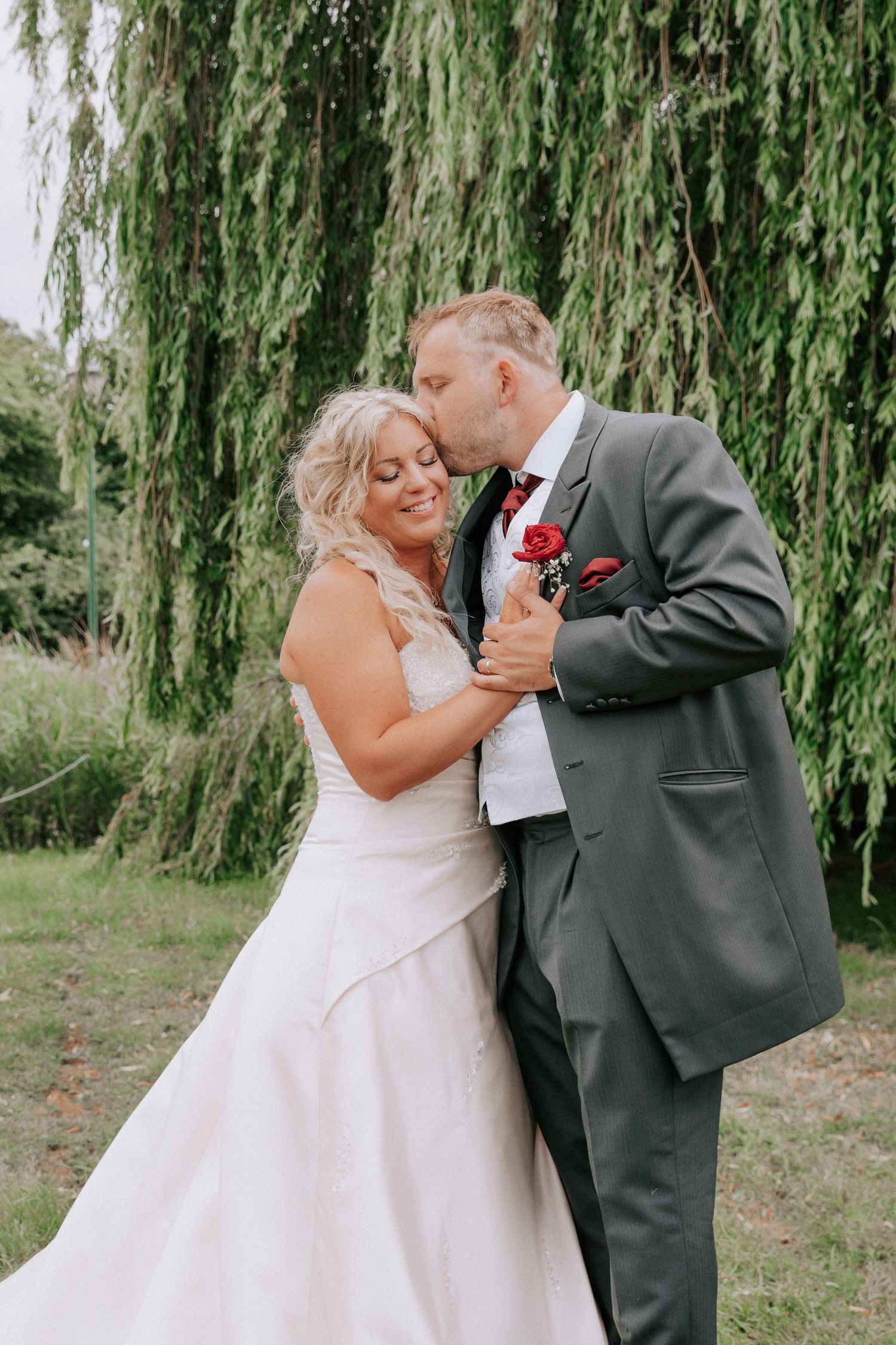 Bride and groom share a romantic kiss under a weeping willow tree, bride in white gown, groom in gray suit.