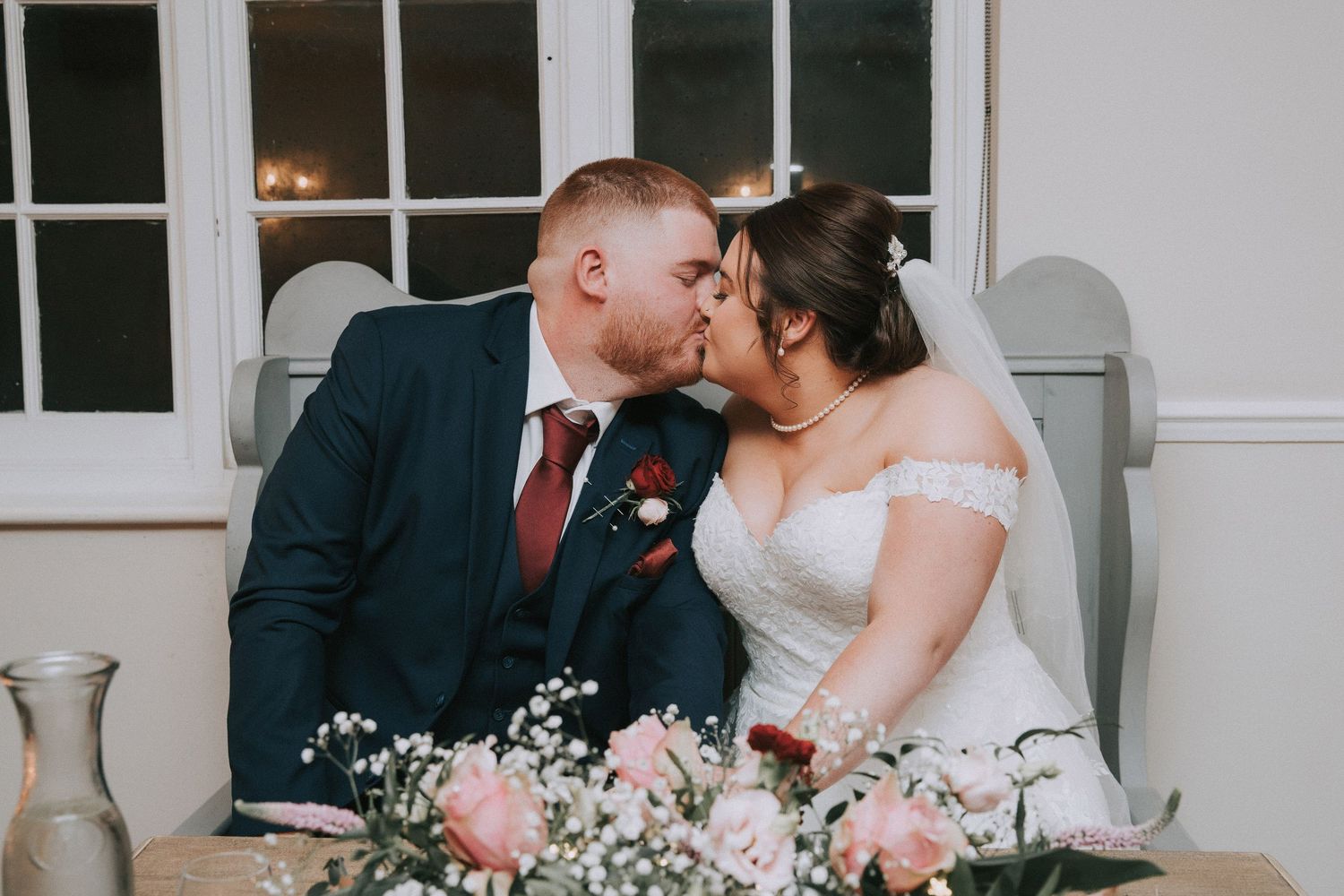Bride and groom share a kiss at wedding reception table decorated with pink roses and baby's breath.
