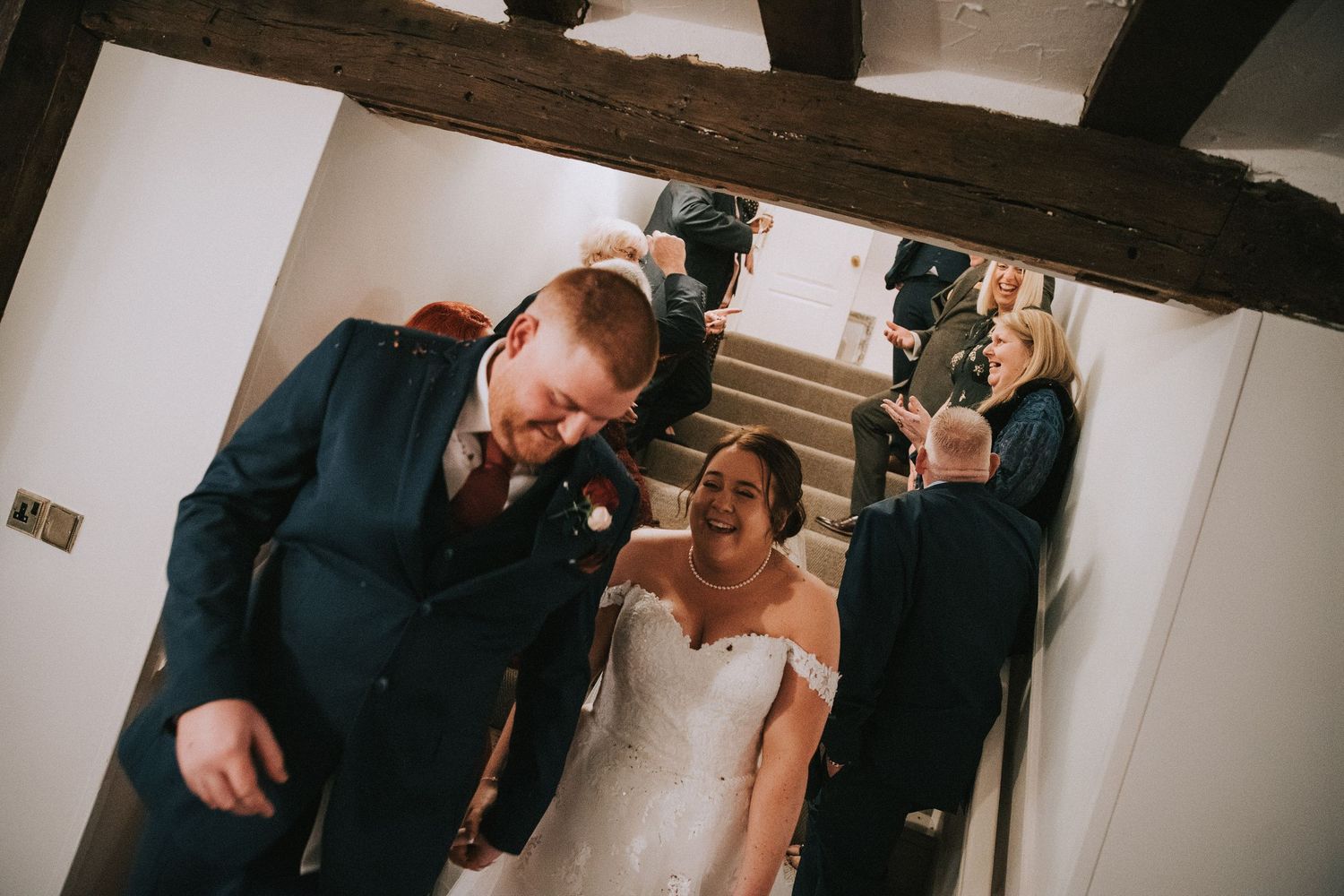 Bride and groom laughing joyfully on staircase at rustic wedding venue with exposed wooden beams.
