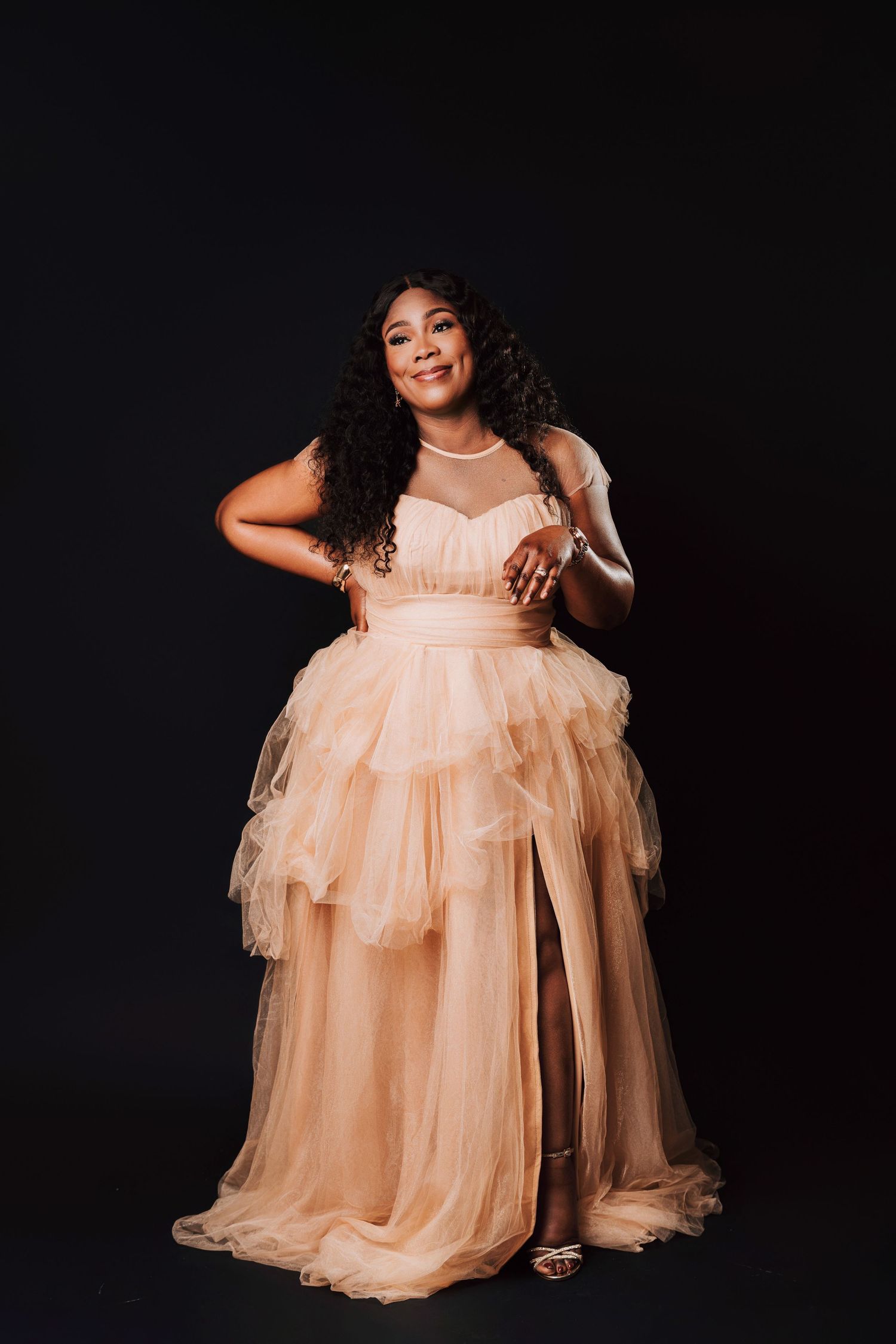 Woman in elegant peach tulle gown posing confidently against black background with curly hair and radiant smile.