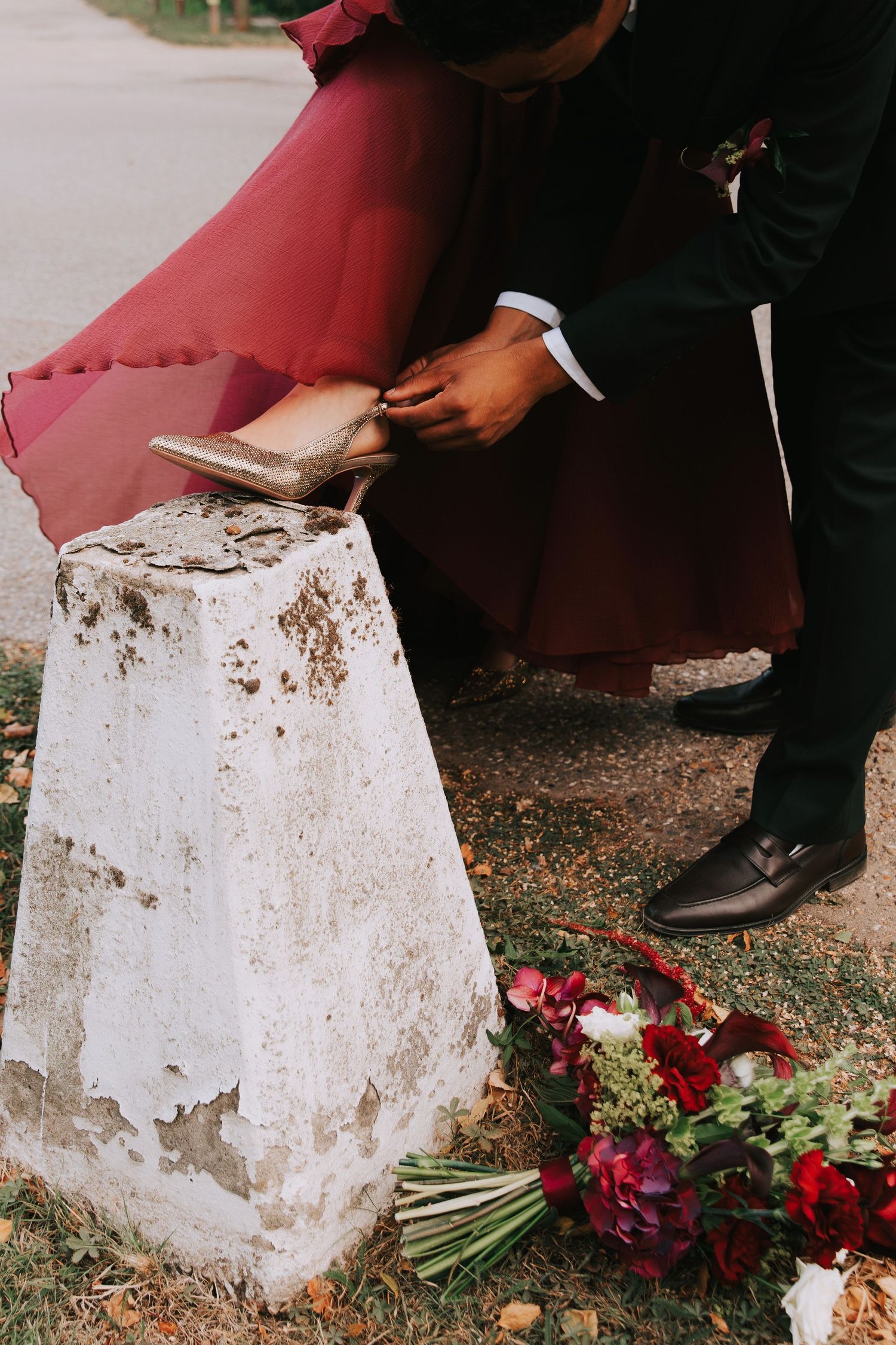 Groom helping bride with gold heels on stone pedestal, red bouquet on ground, wedding couple outdoors.
