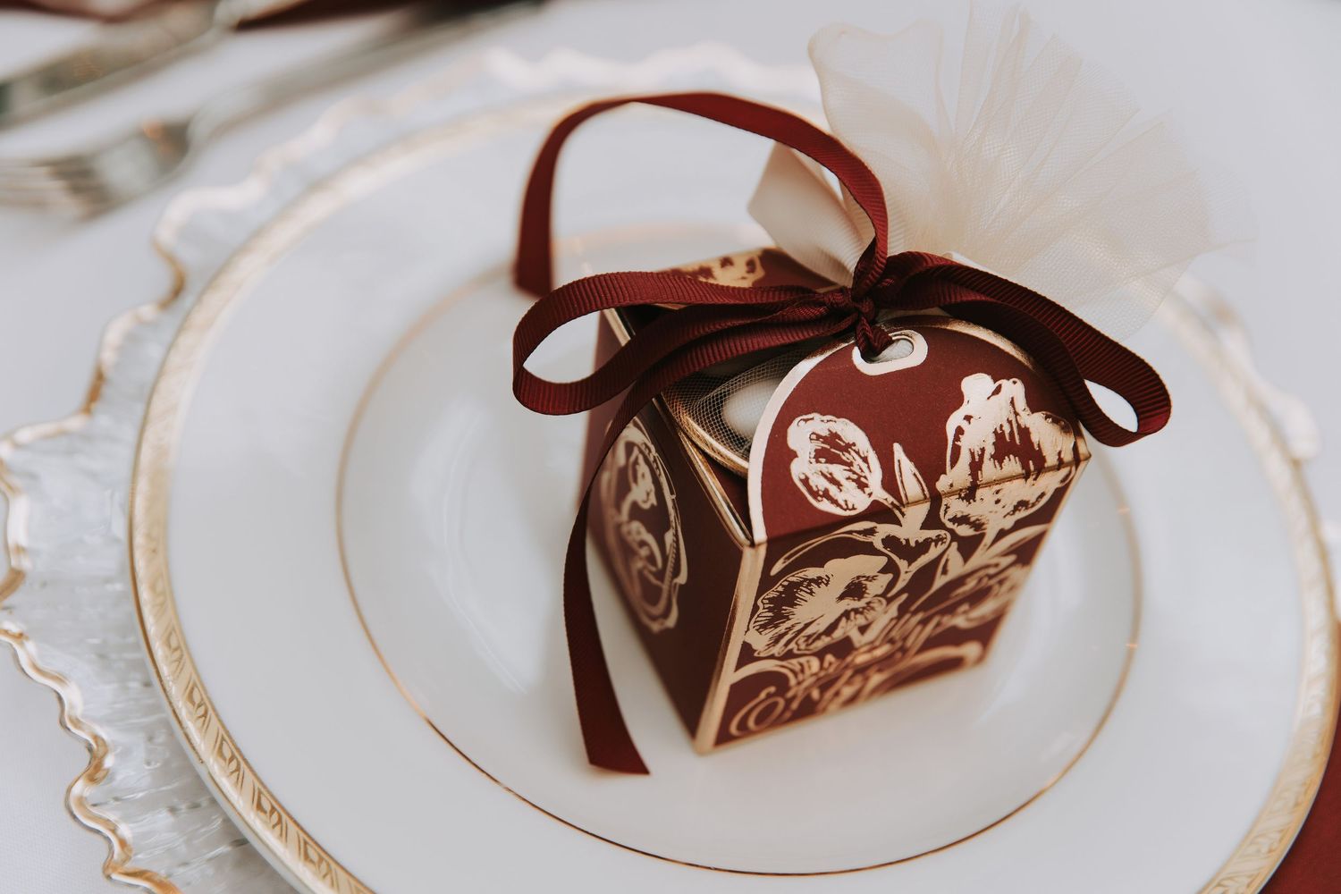 Elegant burgundy and gold wedding favor box with ribbon bow displayed on white china plate at reception table setting.
