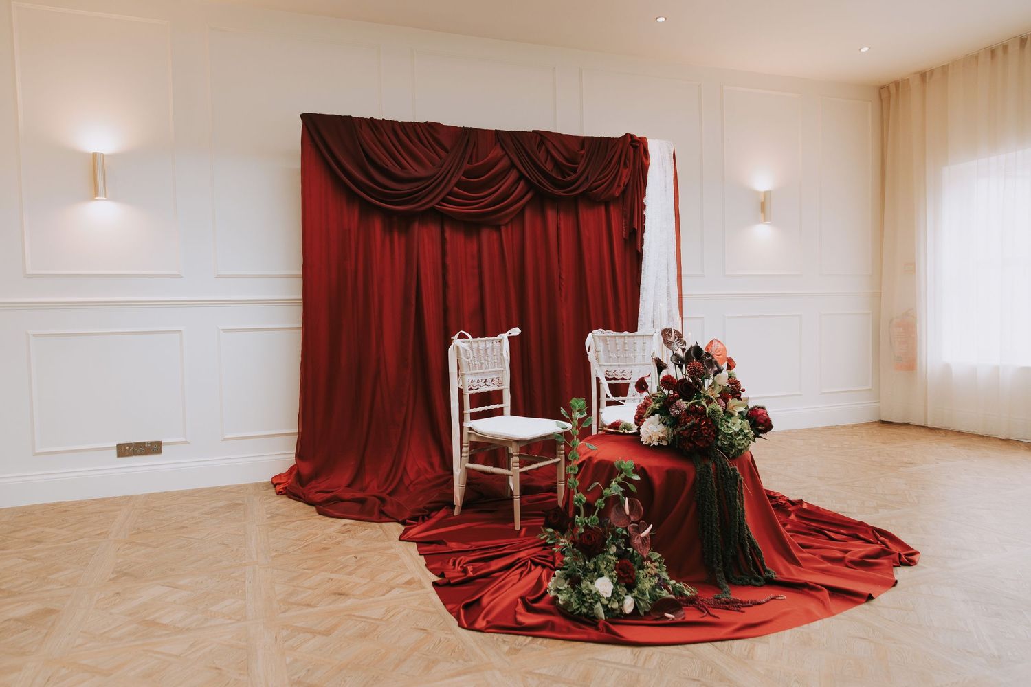 Elegant wedding sweetheart table with white chairs, red velvet draping backdrop, and floral arrangements in a ballroom.