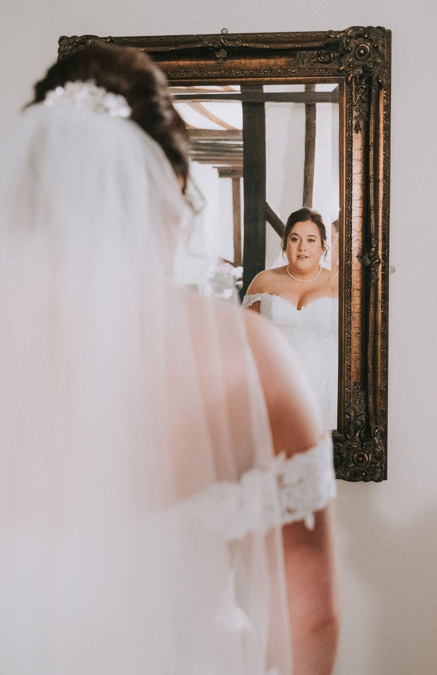 Bride in off-shoulder lace wedding gown and veil admiring her reflection in an ornate gold mirror.