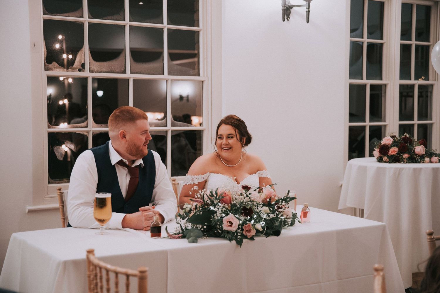 Bride and groom laughing together at sweetheart table with floral centerpiece during wedding reception.