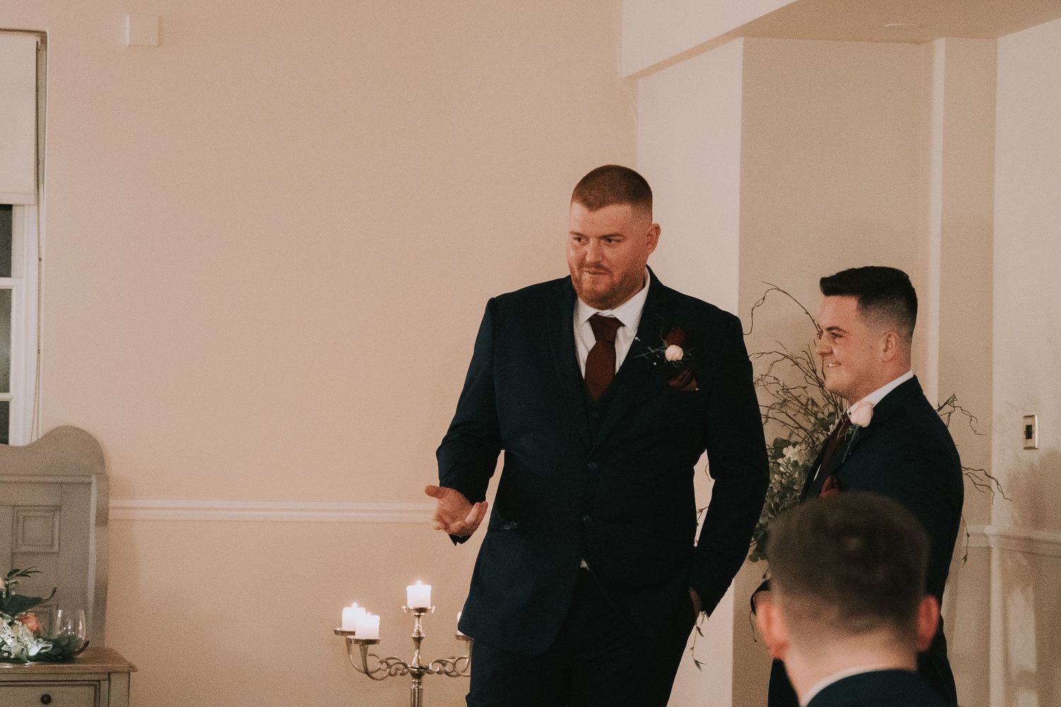 Groom in navy suit with burgundy tie gesturing while standing with groomsmen in a bright wedding venue.