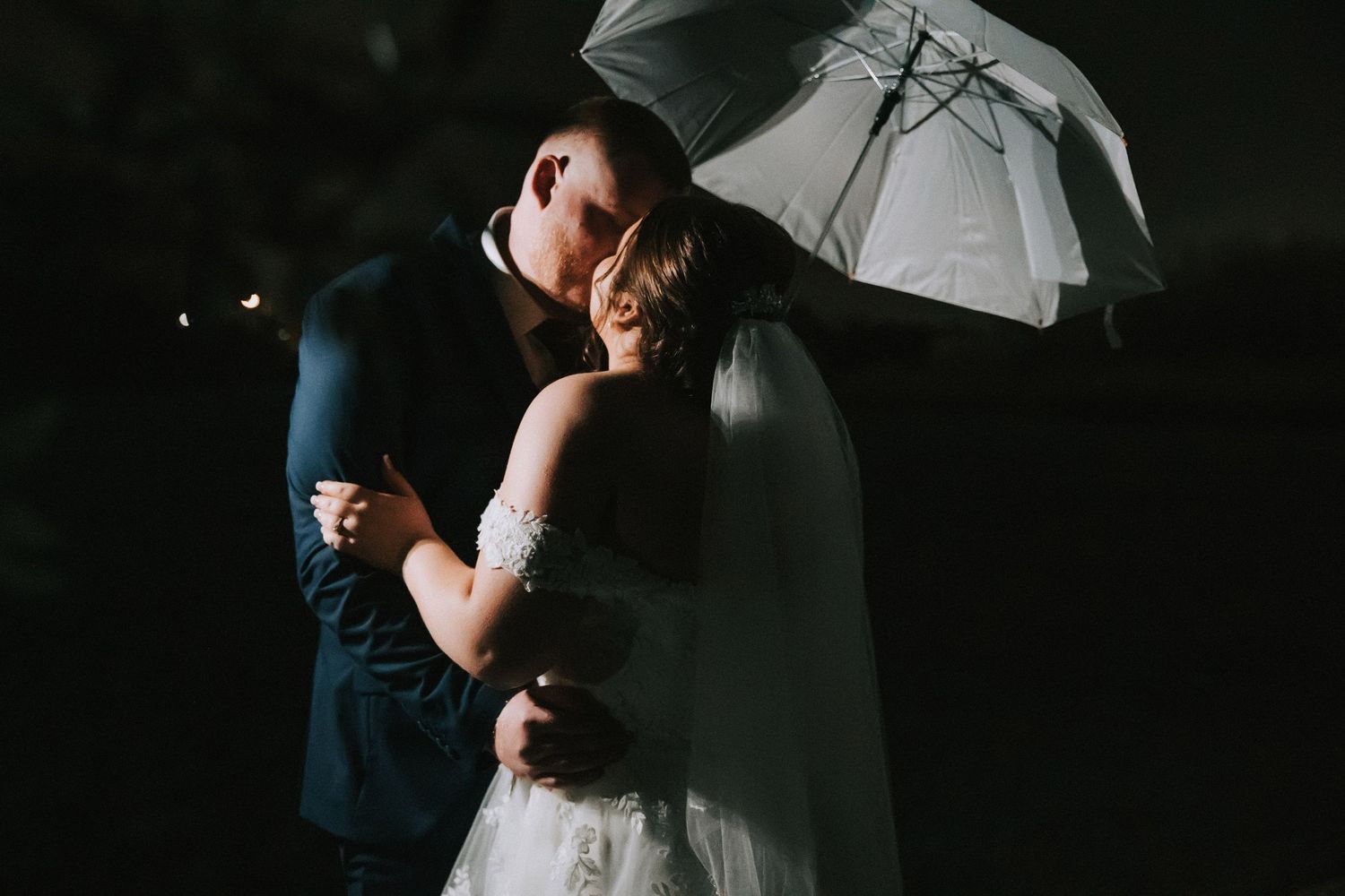 Romantic bride and groom embrace under a white umbrella in a stunning nighttime wedding portrait.