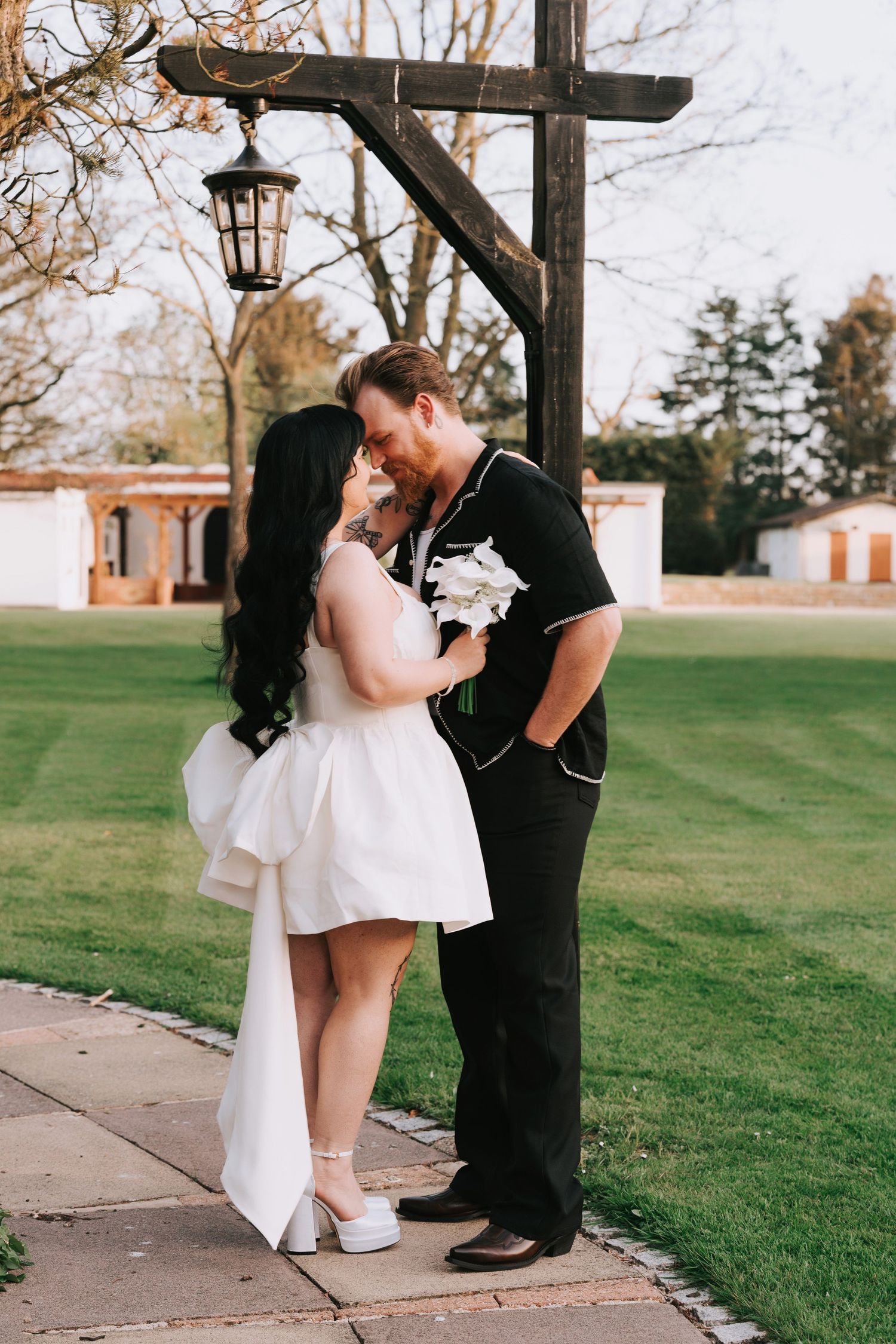 Bride in white mini dress and groom in black suit share romantic moment under wooden lantern post on lush green lawn.
