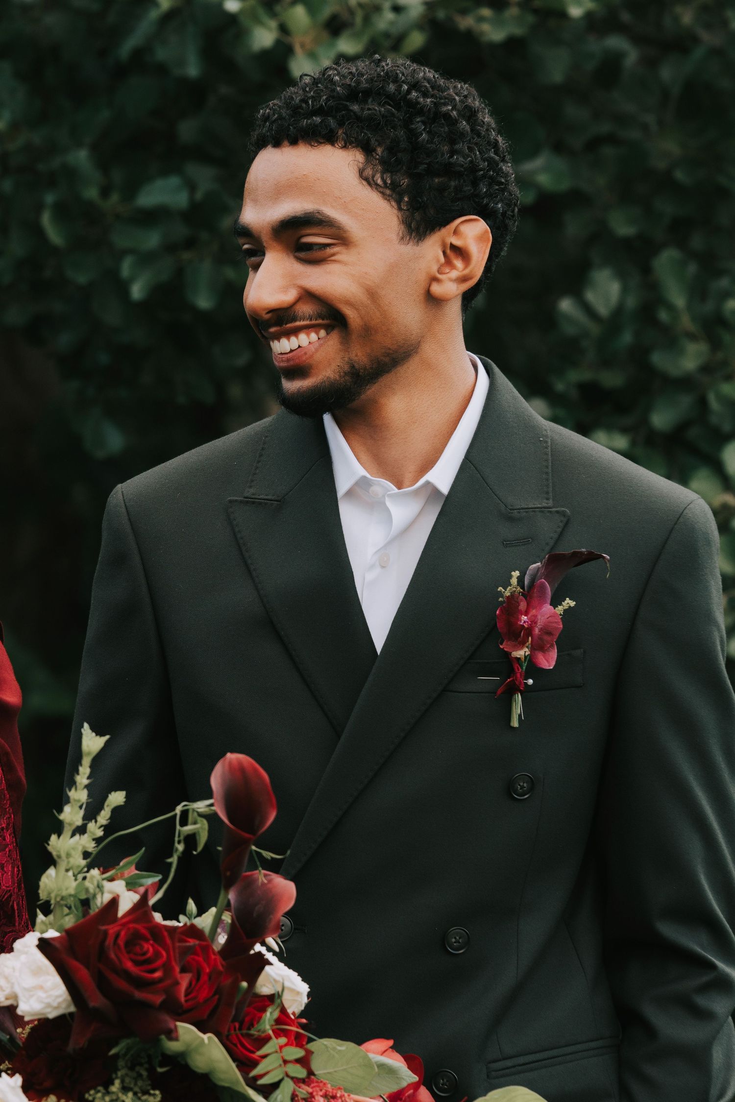 Smiling groom in dark green double-breasted suit with burgundy boutonniere holding red rose bouquet outdoors.