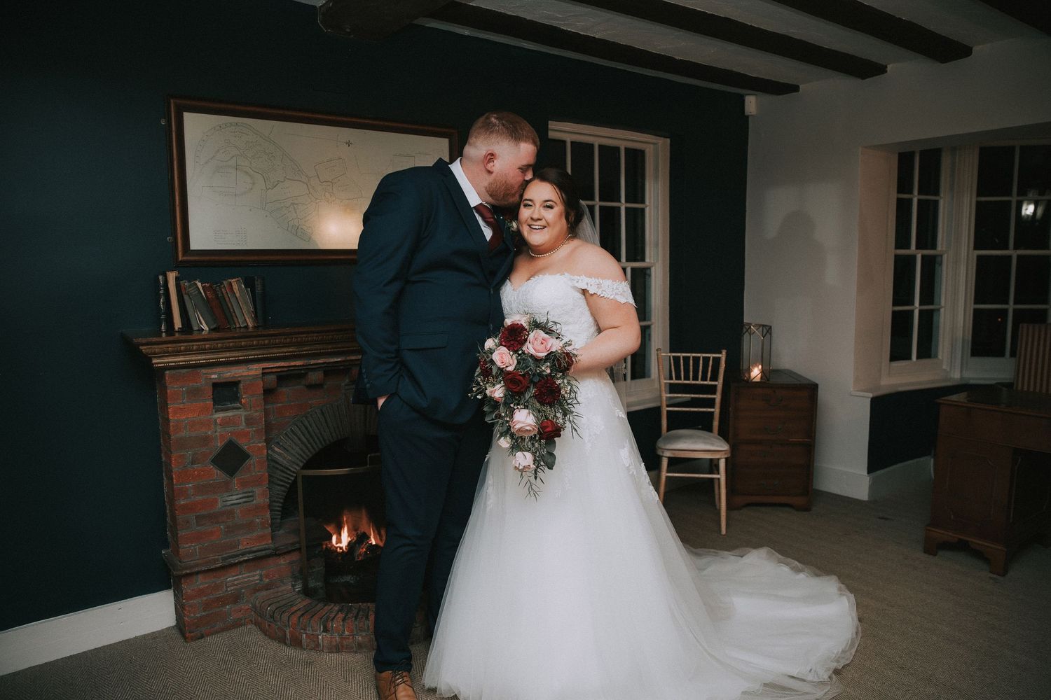 Bride and groom share a romantic moment by a fireplace, bride holding a burgundy bouquet in a white ballgown.
