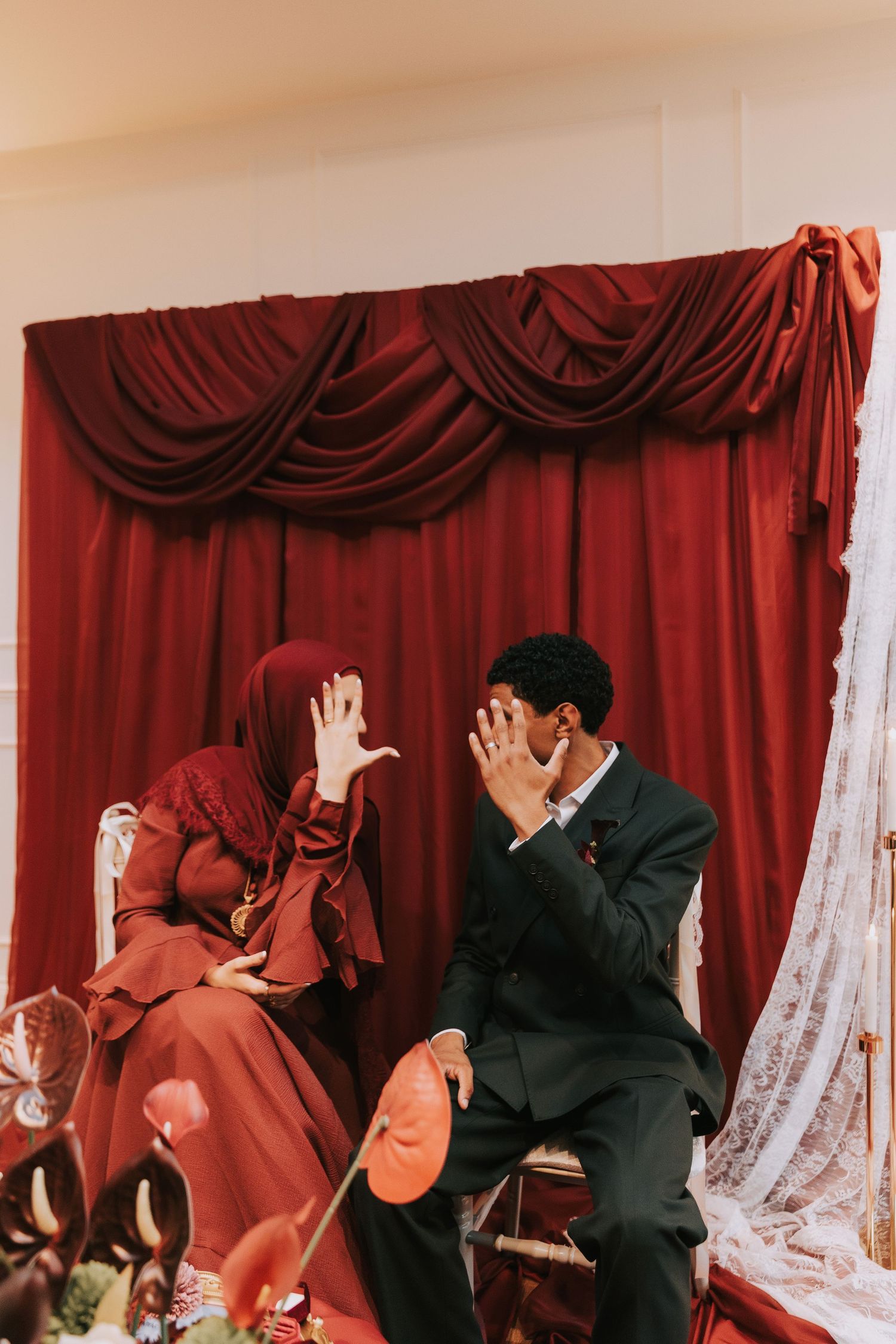Muslim couple in red and black attire sitting together, laughing and covering faces, against elegant red draped curtain backdrop.