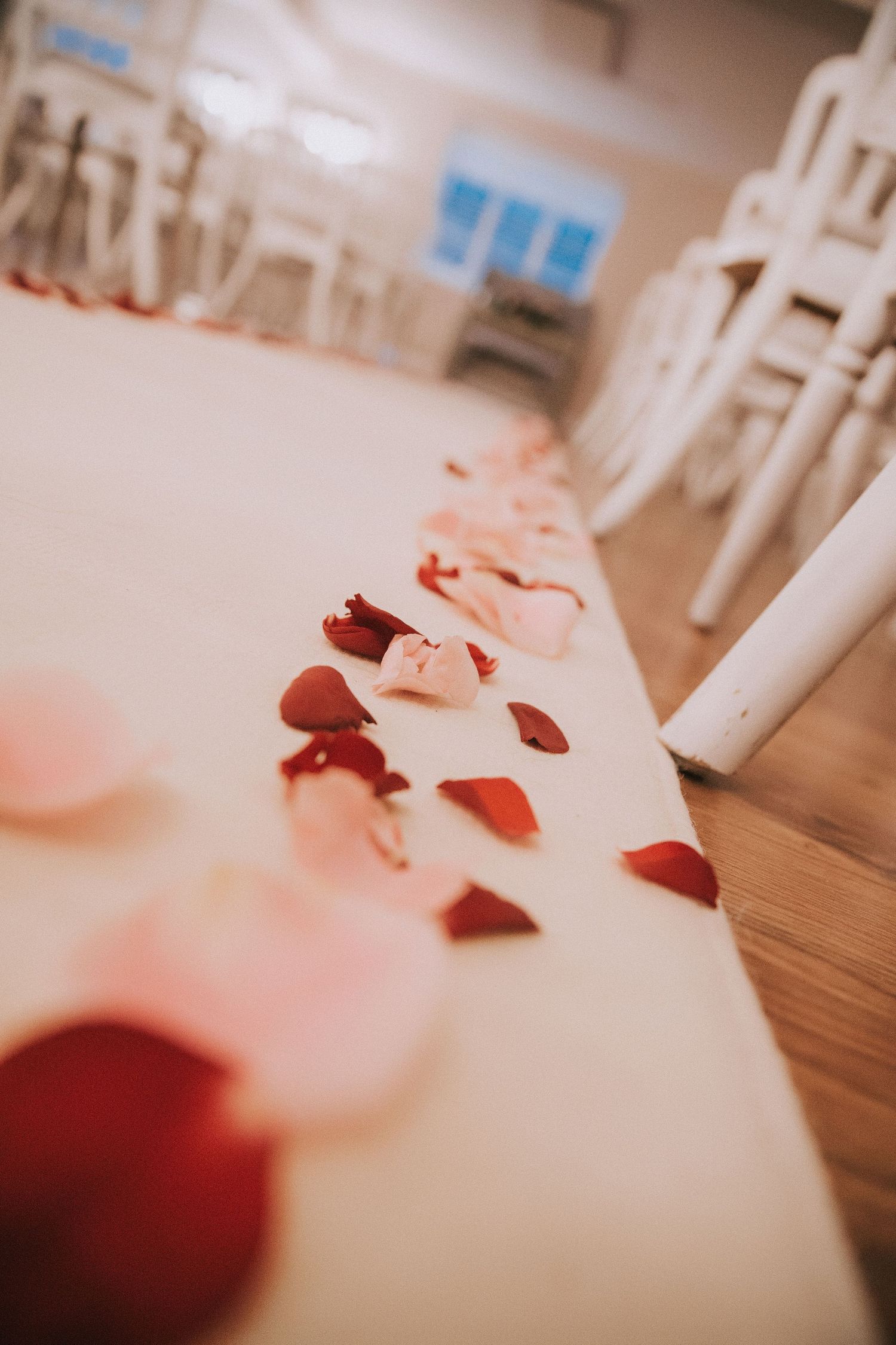 Rose petals scattered along a white wedding aisle runner on wooden floor with white chairs in background.