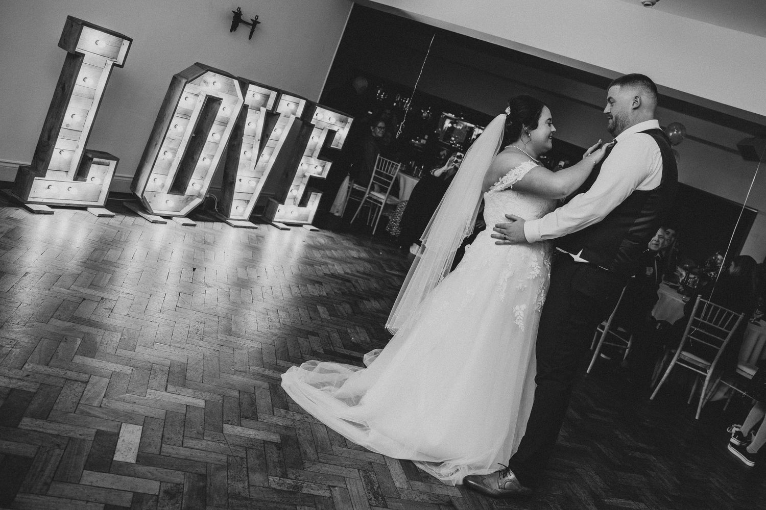 Bride and groom share first dance in black and white photo beside illuminated LOVE letters.
