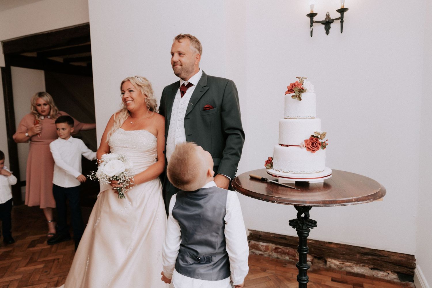 Bride and groom stand near a white wedding cake during their reception ceremony with guests in the background.