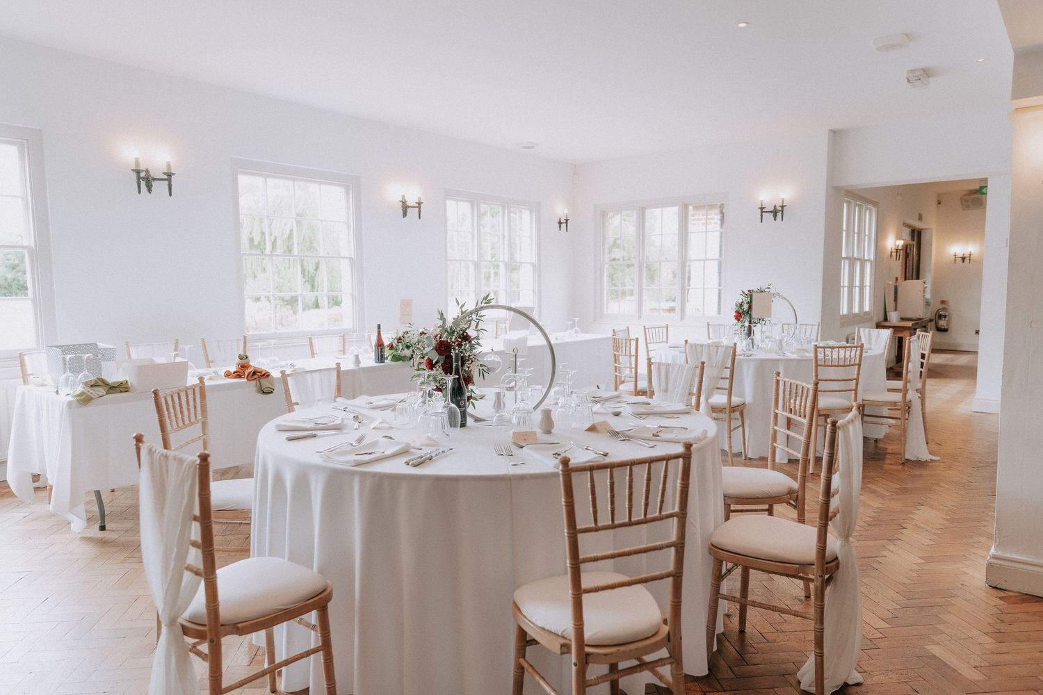 Elegant wedding reception hall with round tables, white linens, gold Chiavari chairs, and herringbone wood floors.