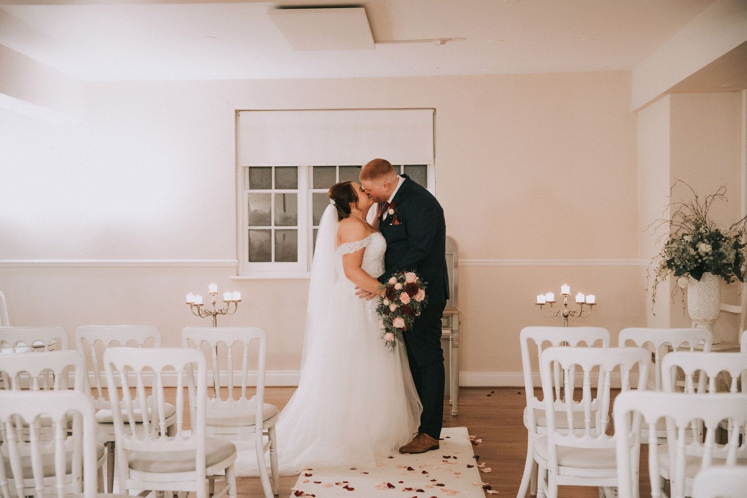 Bride and groom share first kiss at elegant white wedding ceremony venue with white chairs and floral decorations.