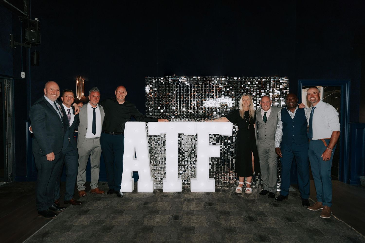 Eight people in formal attire stand behind the company name, set against a shimmering silver backdrop a perfect example of event photography